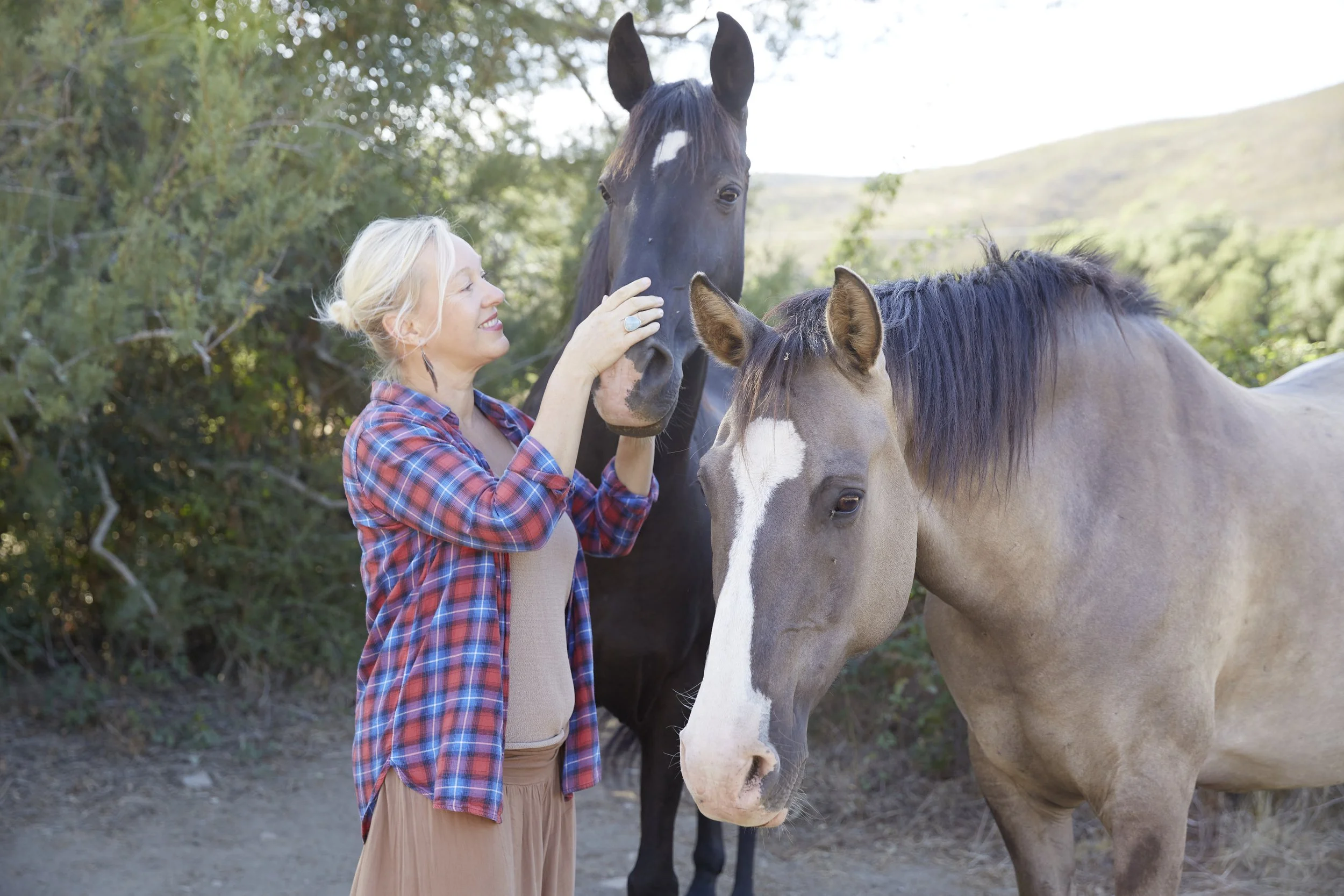 A woman with blonde hair in a ponytail, wearing a red plaid shirt, smiling, petting the nose of a black horse with a white star on its forehead, and a tan horse with a white blaze on its face standing nearby in a wooded outdoor setting.