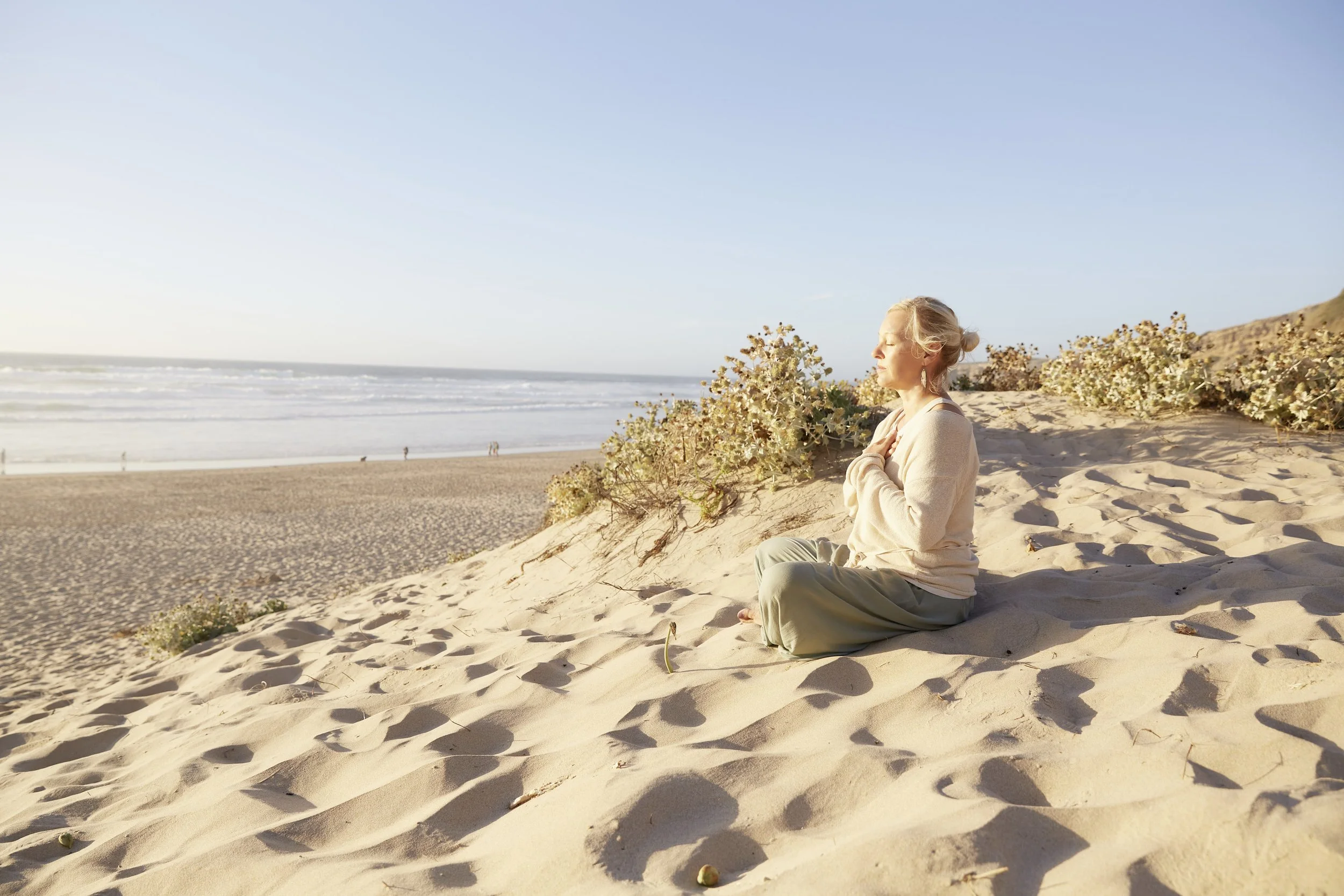 A woman sits cross-legged on sand dunes at the beach, peacefully meditating with her eyes closed, facing the setting sun, with the ocean and a clear blue sky in the background.