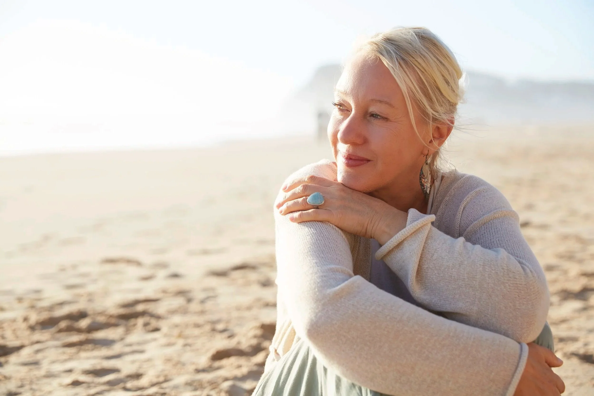 A woman with short blonde hair leaning on her arm, sitting on a sandy beach with the ocean in the background.