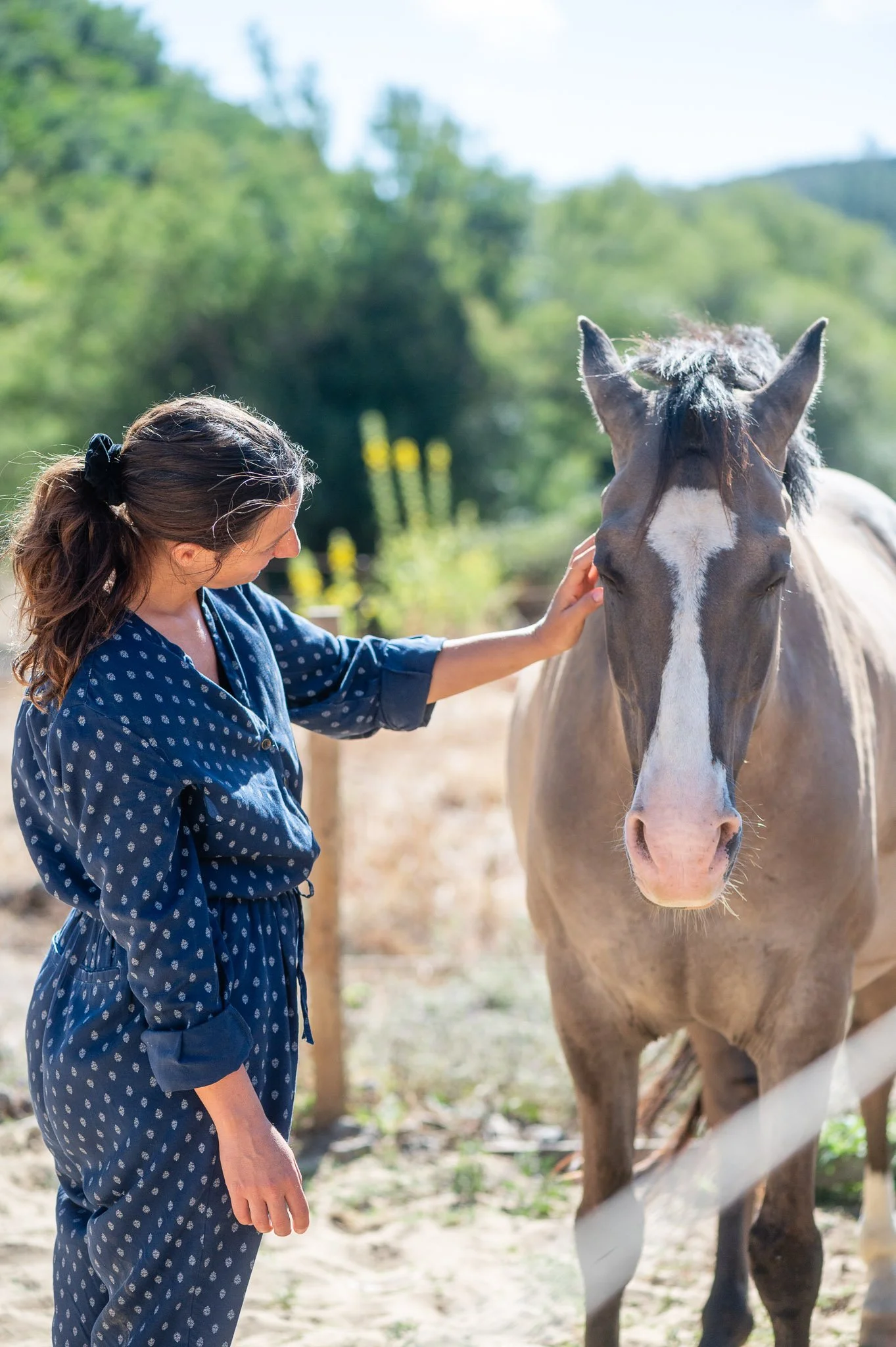 A woman with brown hair tied back, wearing a blue polka dot jumpsuit, gently touching a light brown horse with a white stripe on its face in a sunlit outdoor setting with lush green trees in the background.