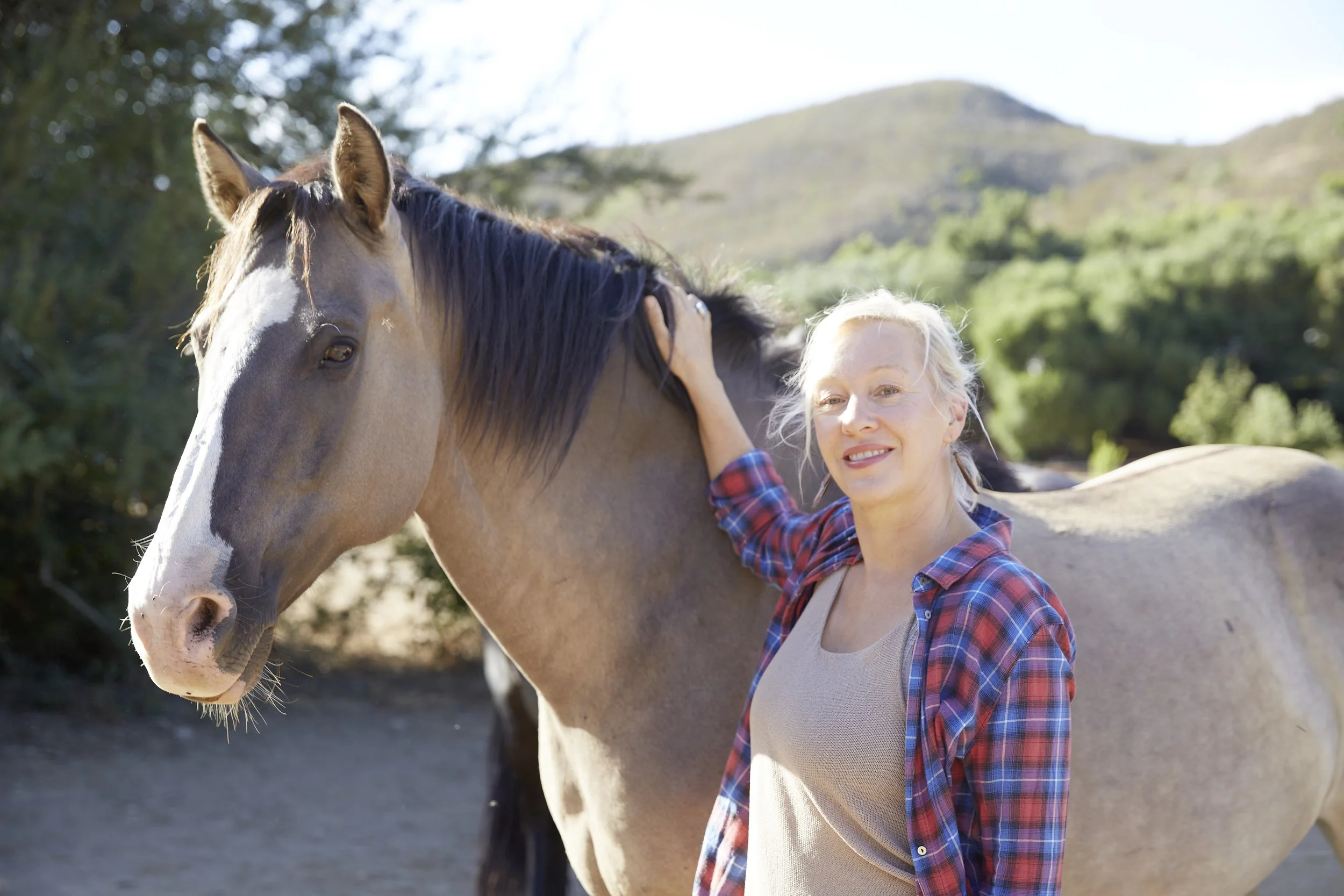 A woman with blonde hair, wearing a beige apron and a plaid shirt, smiling and petting a brown horse with a white face, in an outdoor setting with trees and hills in the background.
