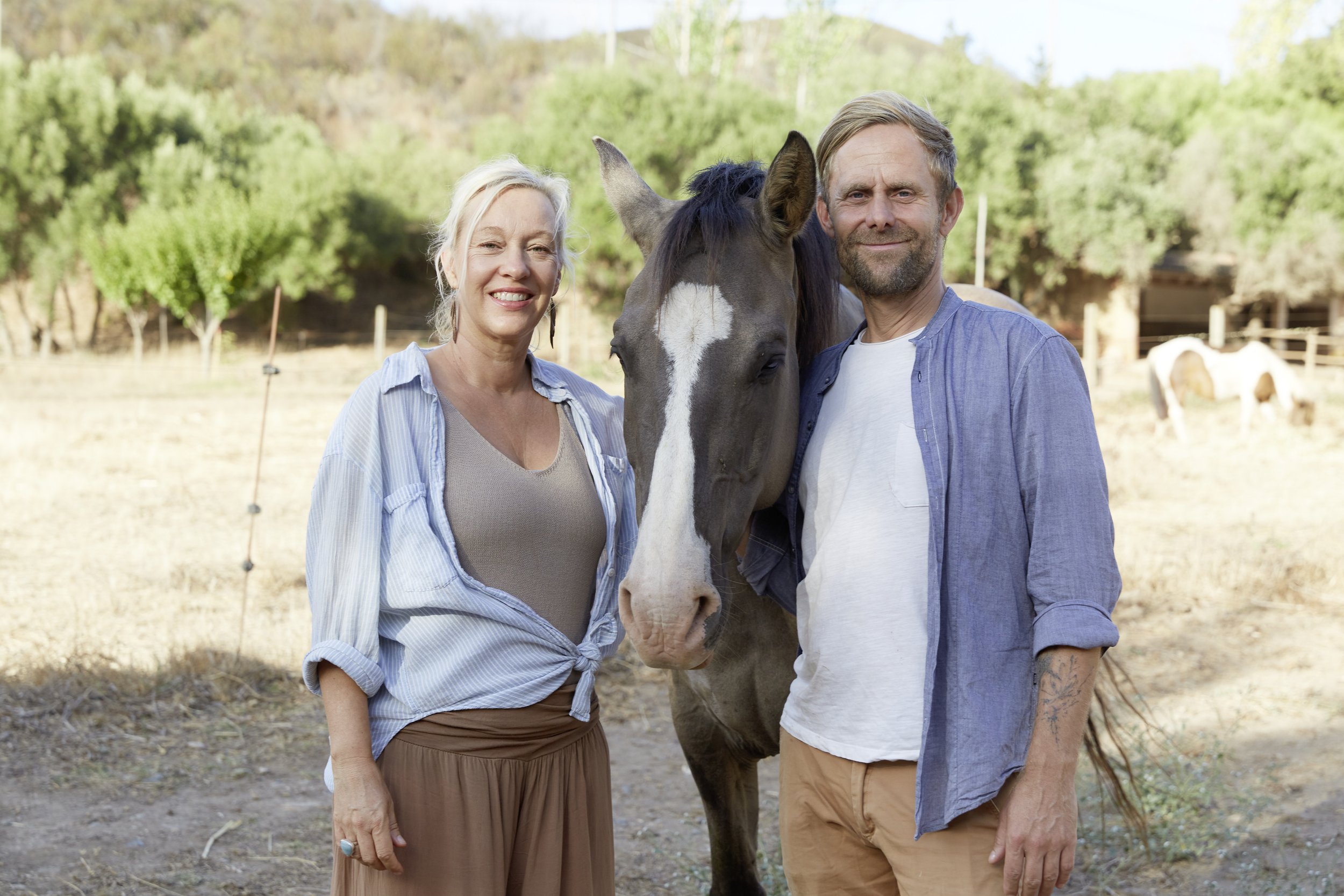 A woman and a man standing outdoors with a horse between them, smiling at the camera, in a natural setting with trees and dry grass.