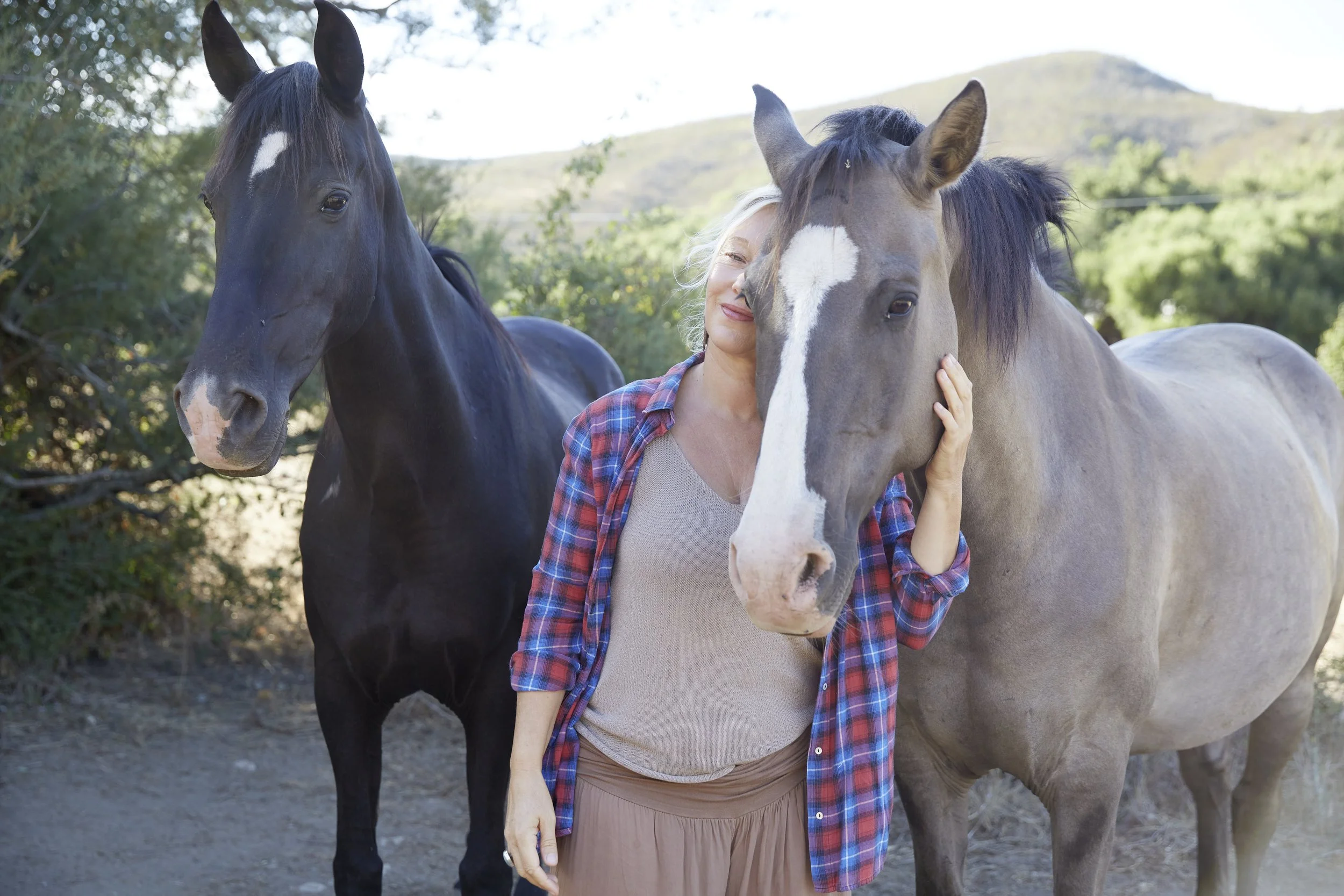 A woman with blonde hair and a plaid shirt hugging a gray horse with a white face mark, standing outdoors with another black horse nearby, trees, and hills in the background.