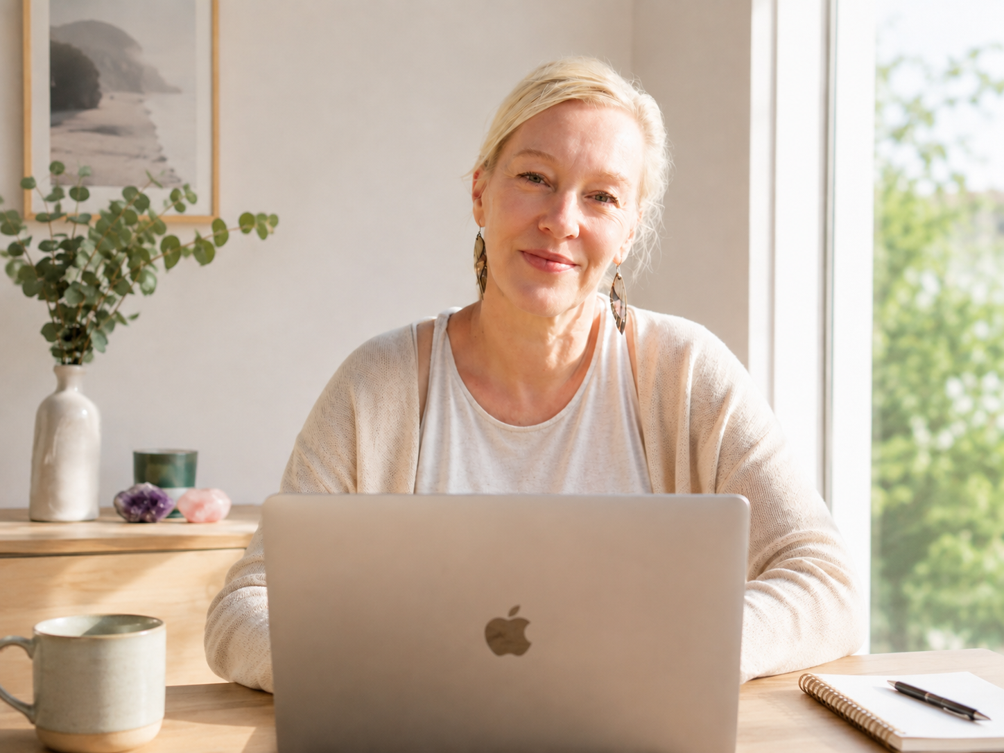 A woman with short blond hair smiling at a laptop at a wooden desk by a large window with a green outdoor view. The desk has a notebook, pen, and a ceramic mug. In the background, there is a white vase with green foliage, a framed beach landscape, and crystals on a shelf.
