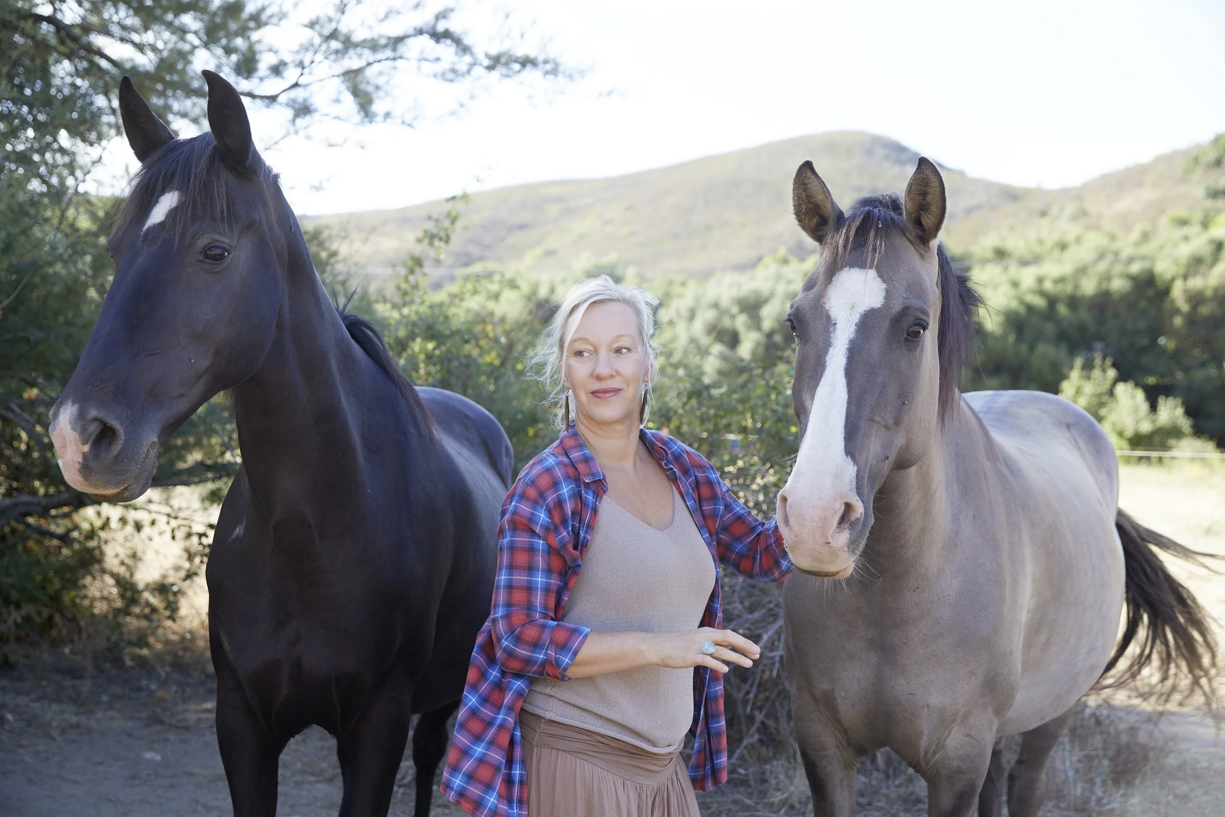 A woman with gray hair, wearing a plaid shirt and a light-colored dress, standing between two horses outdoors in a natural setting with hills and trees in the background.