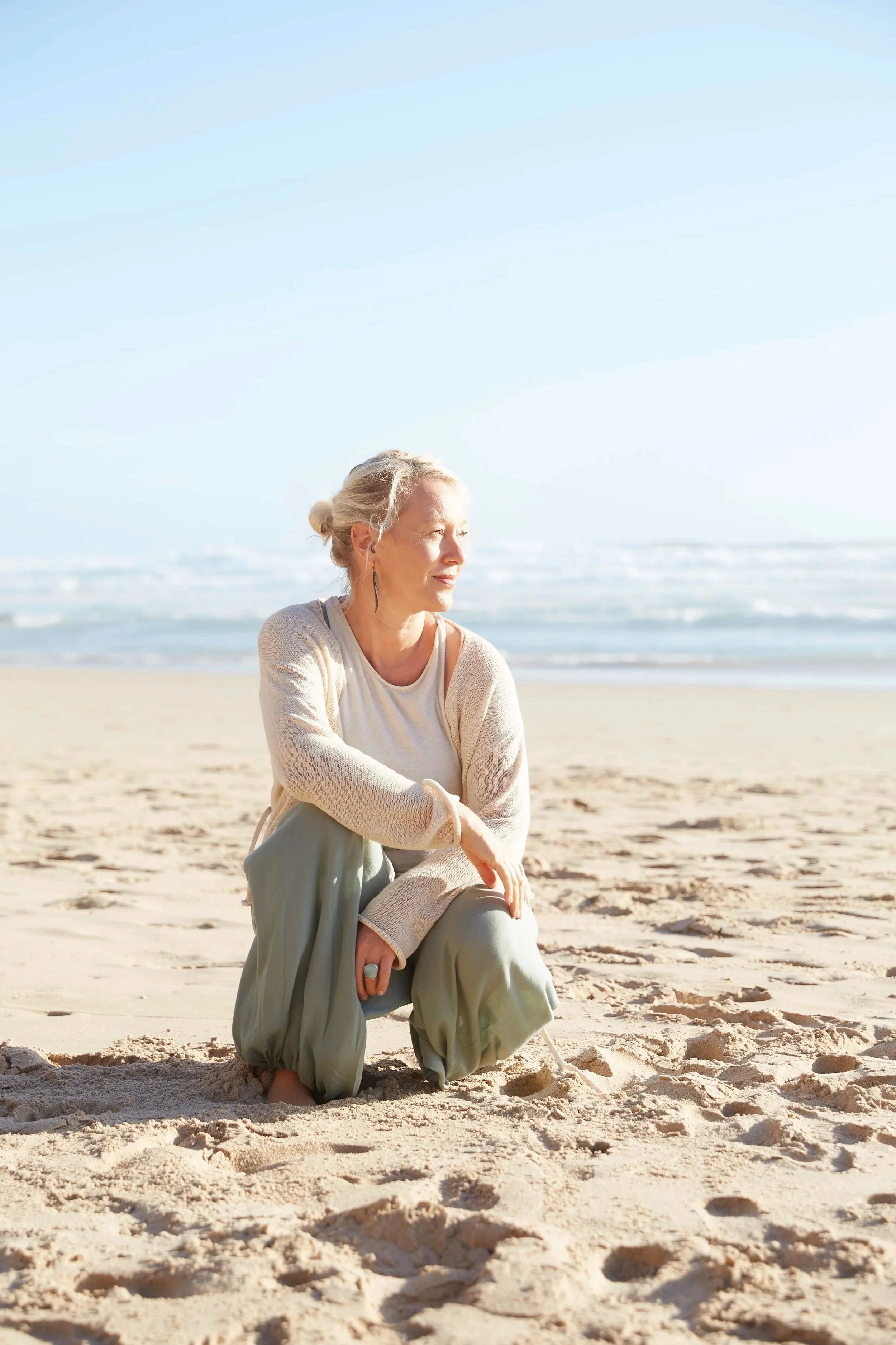 A woman with gray hair in a bun sitting on the sand at the beach during daytime, with the ocean and clear sky in the background.