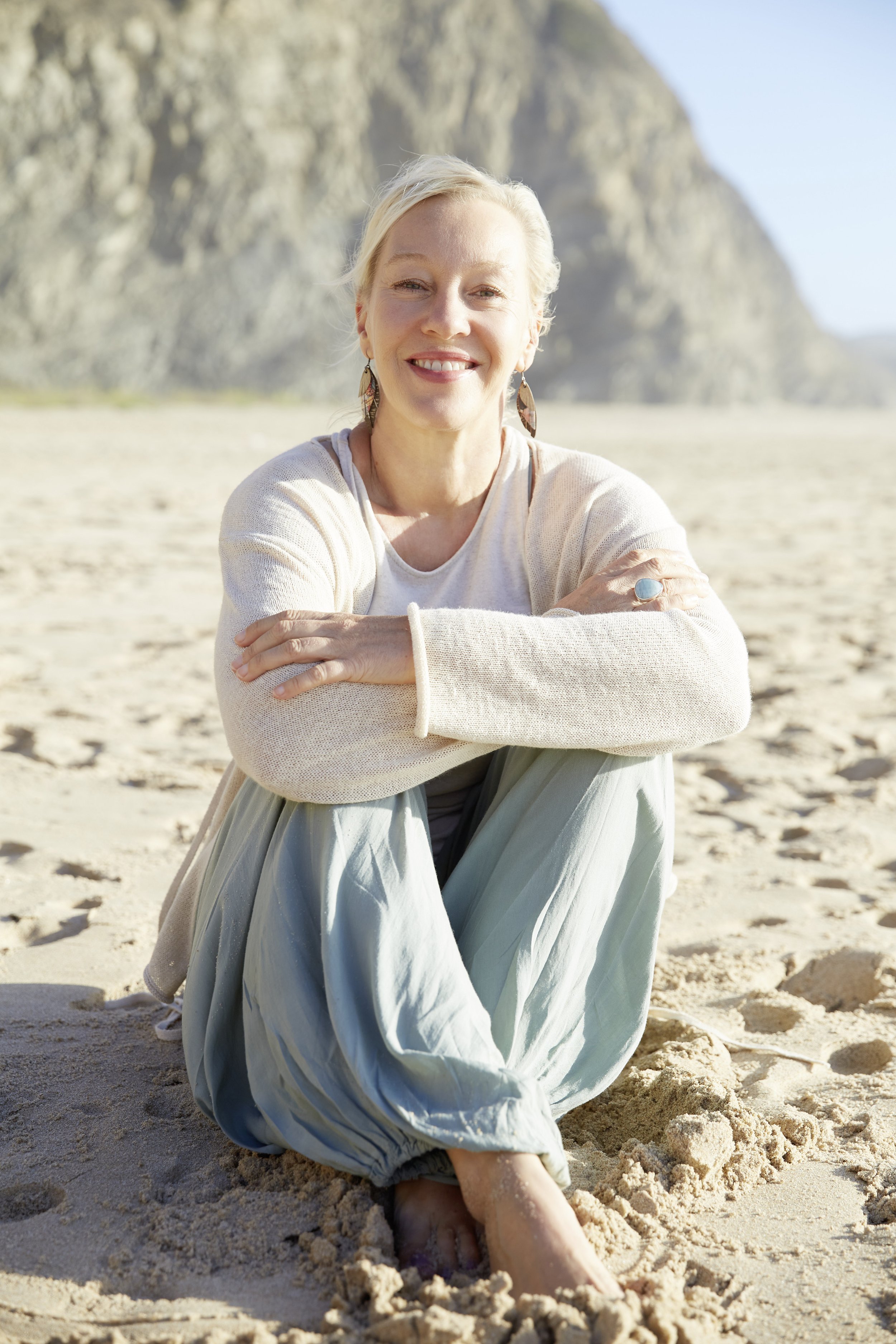 A woman with short blond hair smiling, sitting on a sandy beach with her legs crossed. She is wearing a light-colored sweater, a large ring, big earrings, and loose pants. Behind her are rocky cliffs under a clear blue sky.
