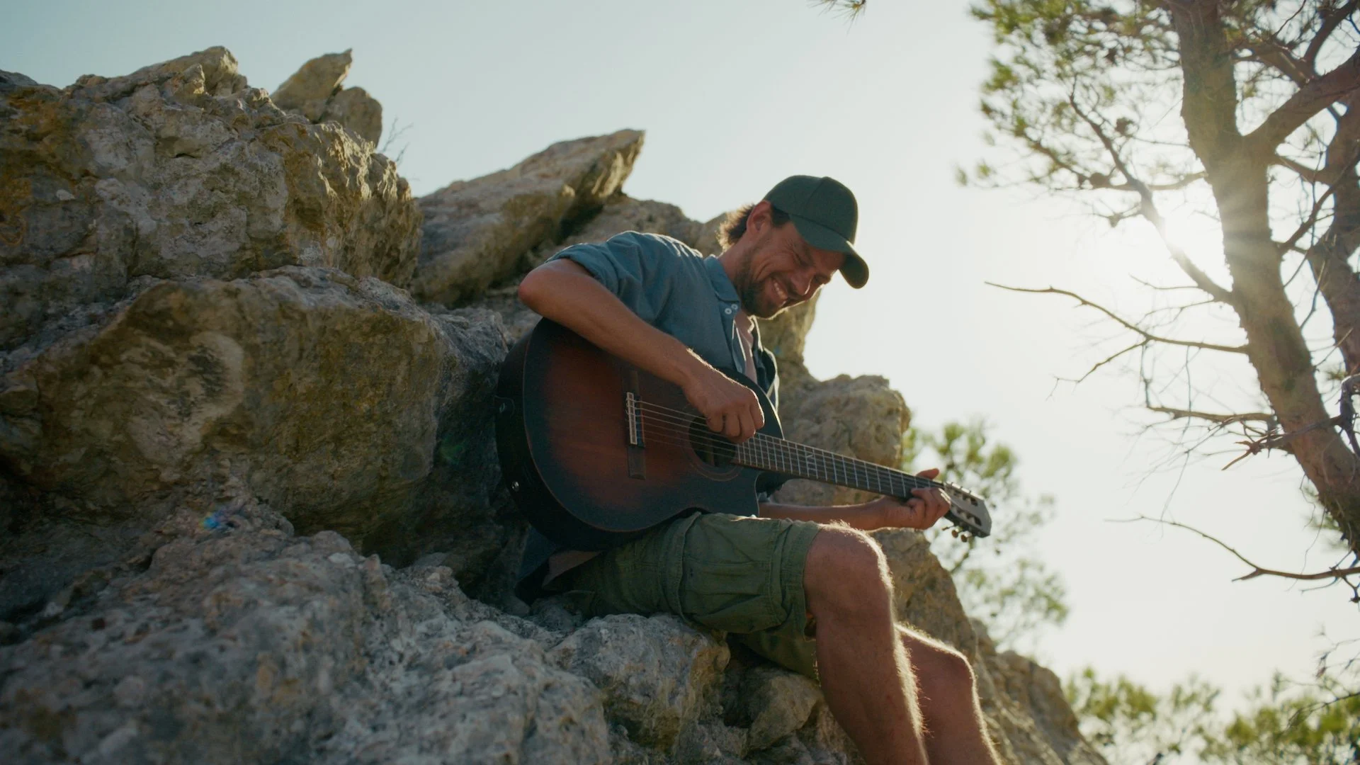 Samuel Boyer spielt Gitarre auf einem Felsen im Freien bei Sonnenlicht, umgeben von Natur, mit einem Baum im Hintergrund. (Mallorca Solar Experience) Foto: Benjamin Sternad