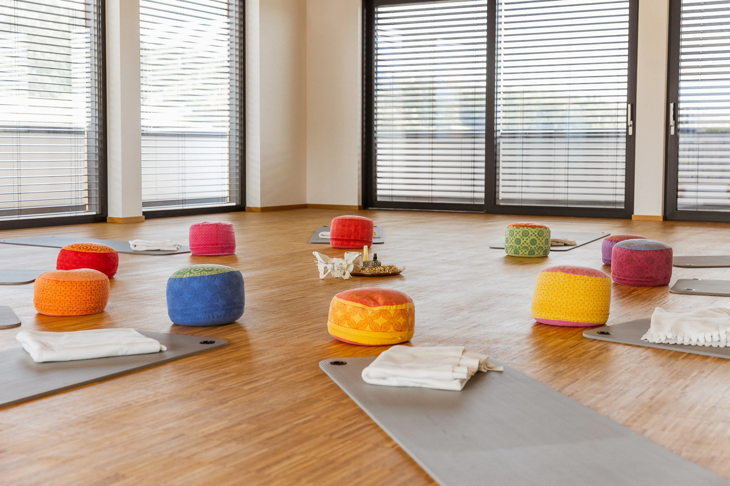 Ein Yoga- oder Meditationsraum mit bunten Sitzkissen, Yogamatten, weißen Handtüchern, einem Schädel, einem Räucherstäbchenhalter und einem kleinen Duftöl. Bei Tageslicht durch große Fenster mit geschlossenen Jalousien.