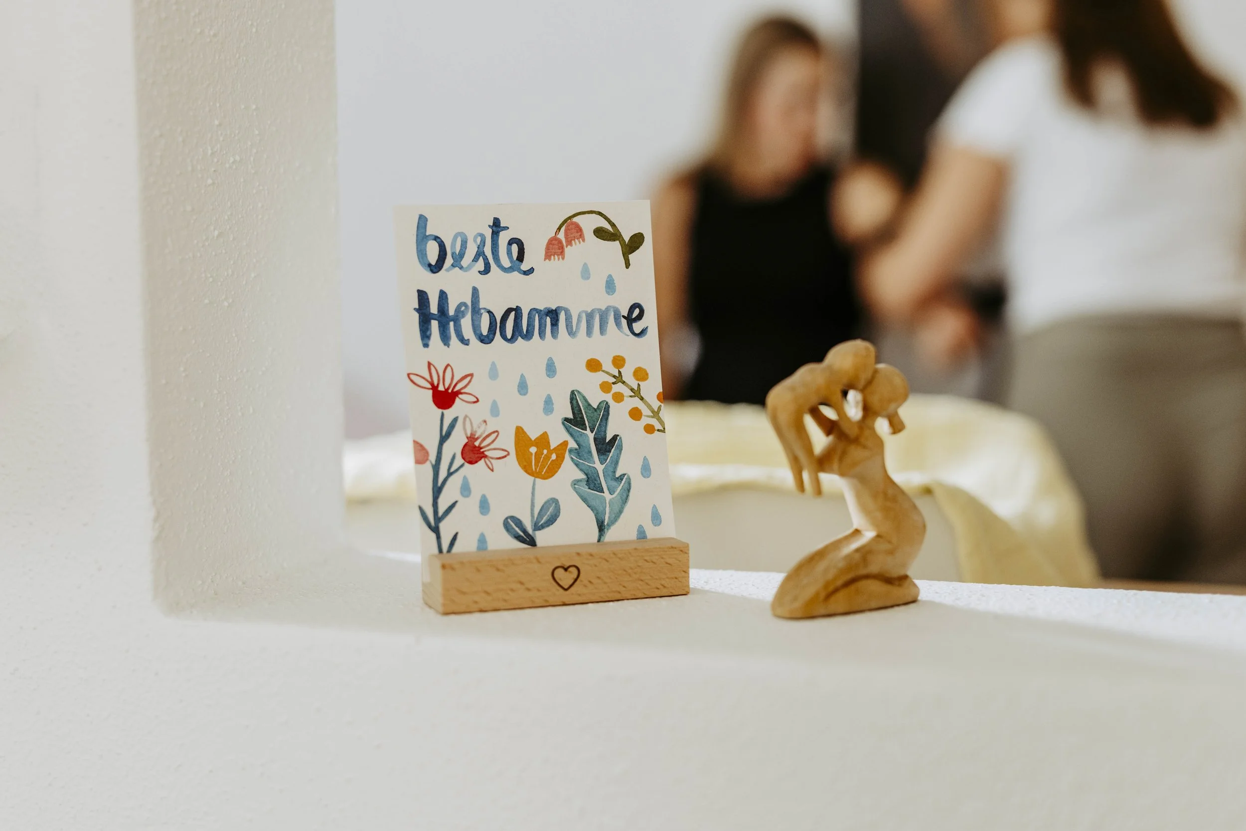 Ein handgeschriebener Glückwunschkarte mit der Aufschrift: "Beste Hebamme" und bunten Blumen, auf einem Holzständer.