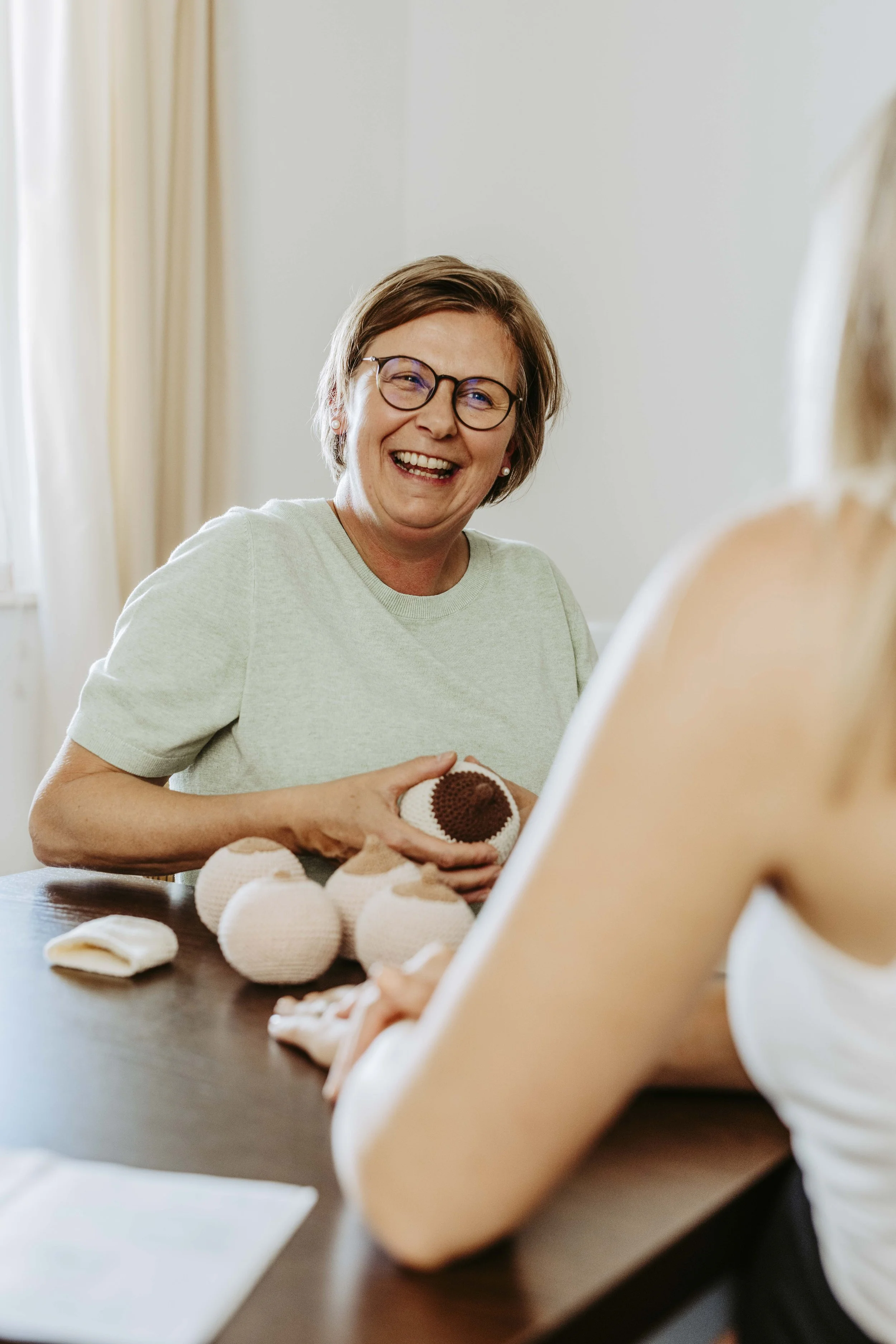 Eine lachende Frau mit Brille und Kurzhaarfrisur sitzt an einem Tisch, hält ein gestricktes Modell in den Händen und spricht mit einer Person, die im Vordergrund zu sehen ist.