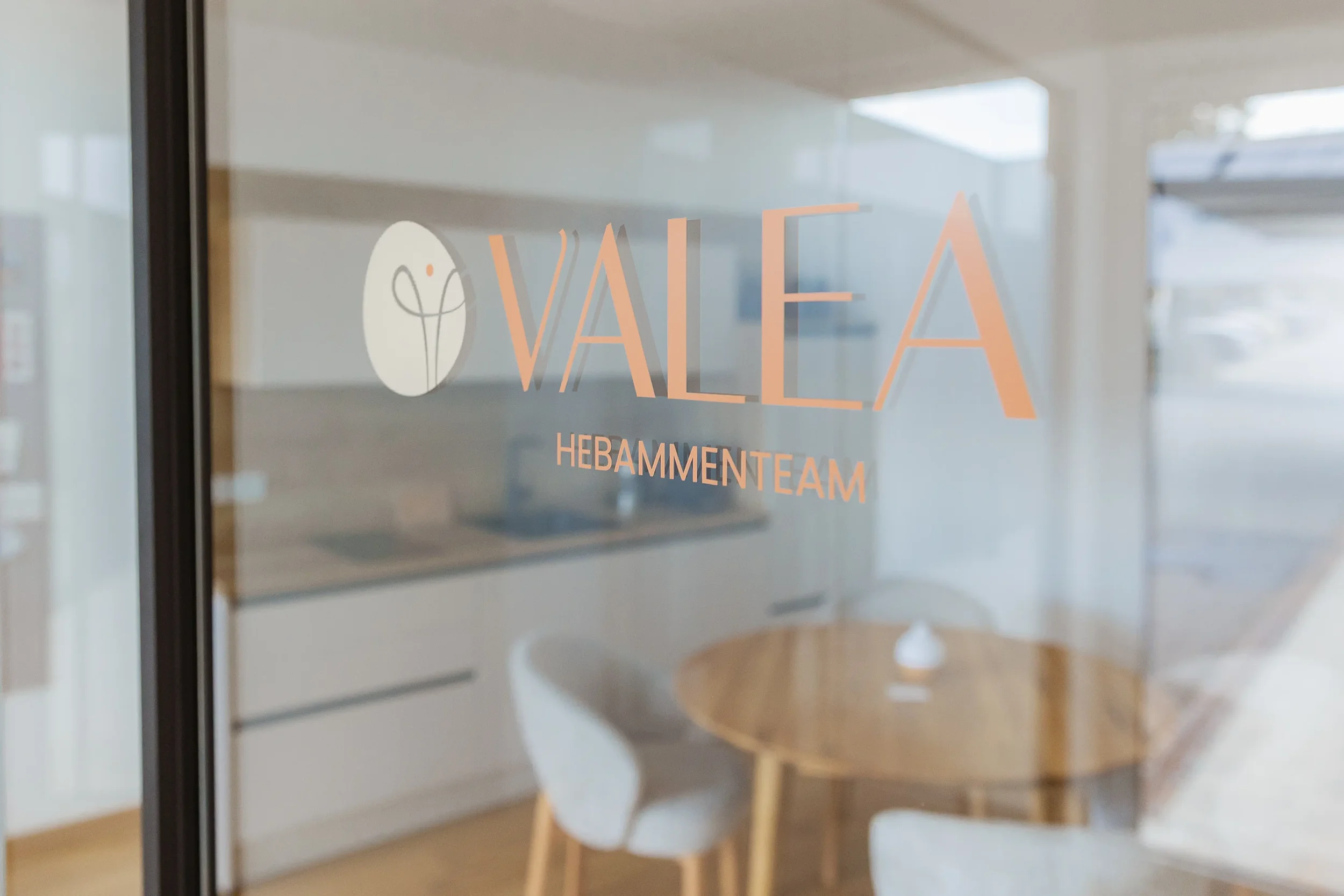 Blick durch eine gläserne Tür mit einem Logo und dem Text 'VALEA Hebammenteam'. Innen sind helle Wände, ein Tisch und Stühle sichtbar.