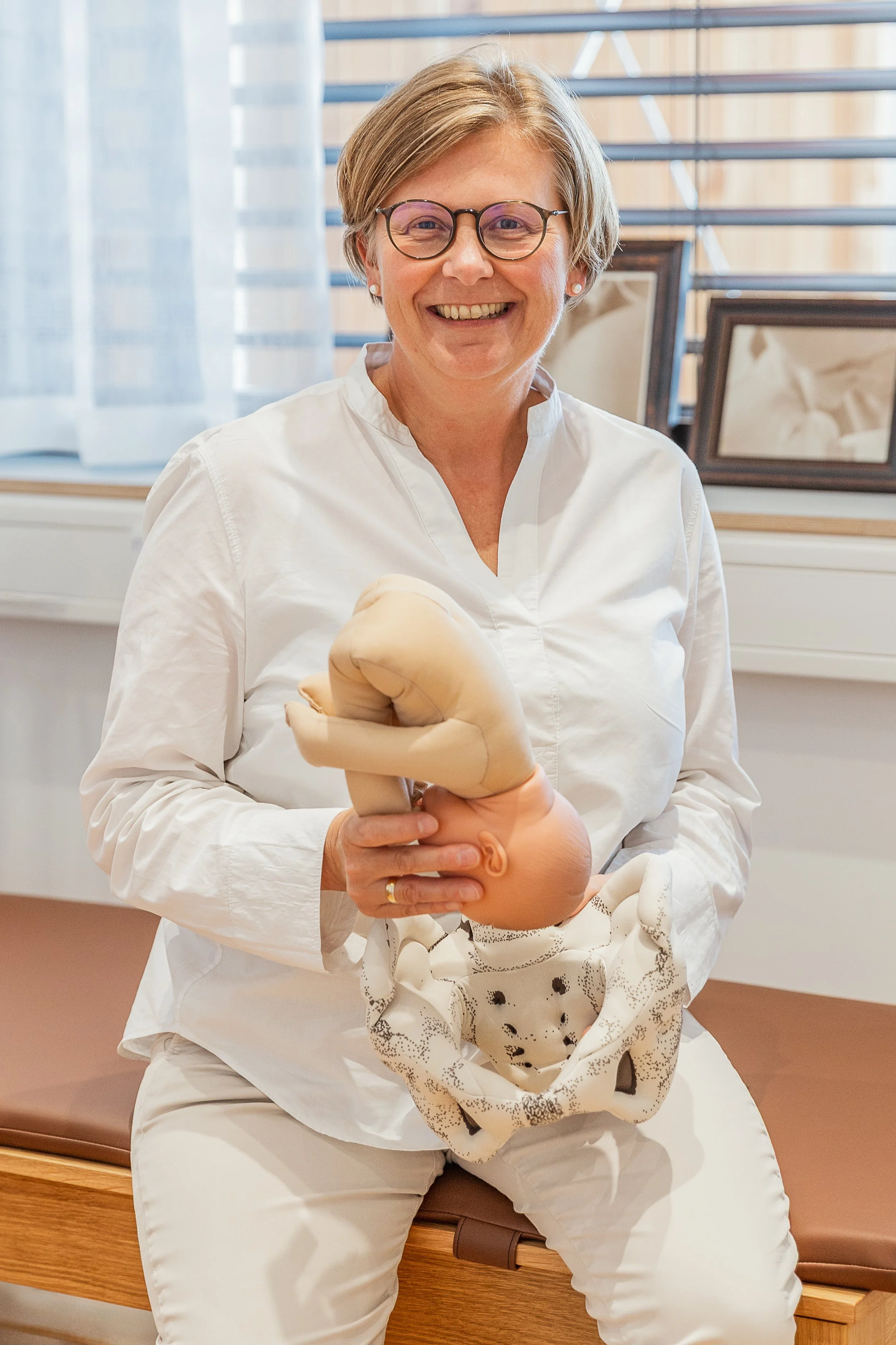 Eine Frau mit Kurzhaar, Brille und weißen Kittel, die eine Baby-Puppe hält, sitzt auf einer Bank in einem Raum mit großen Fenstern und Vorhängen.