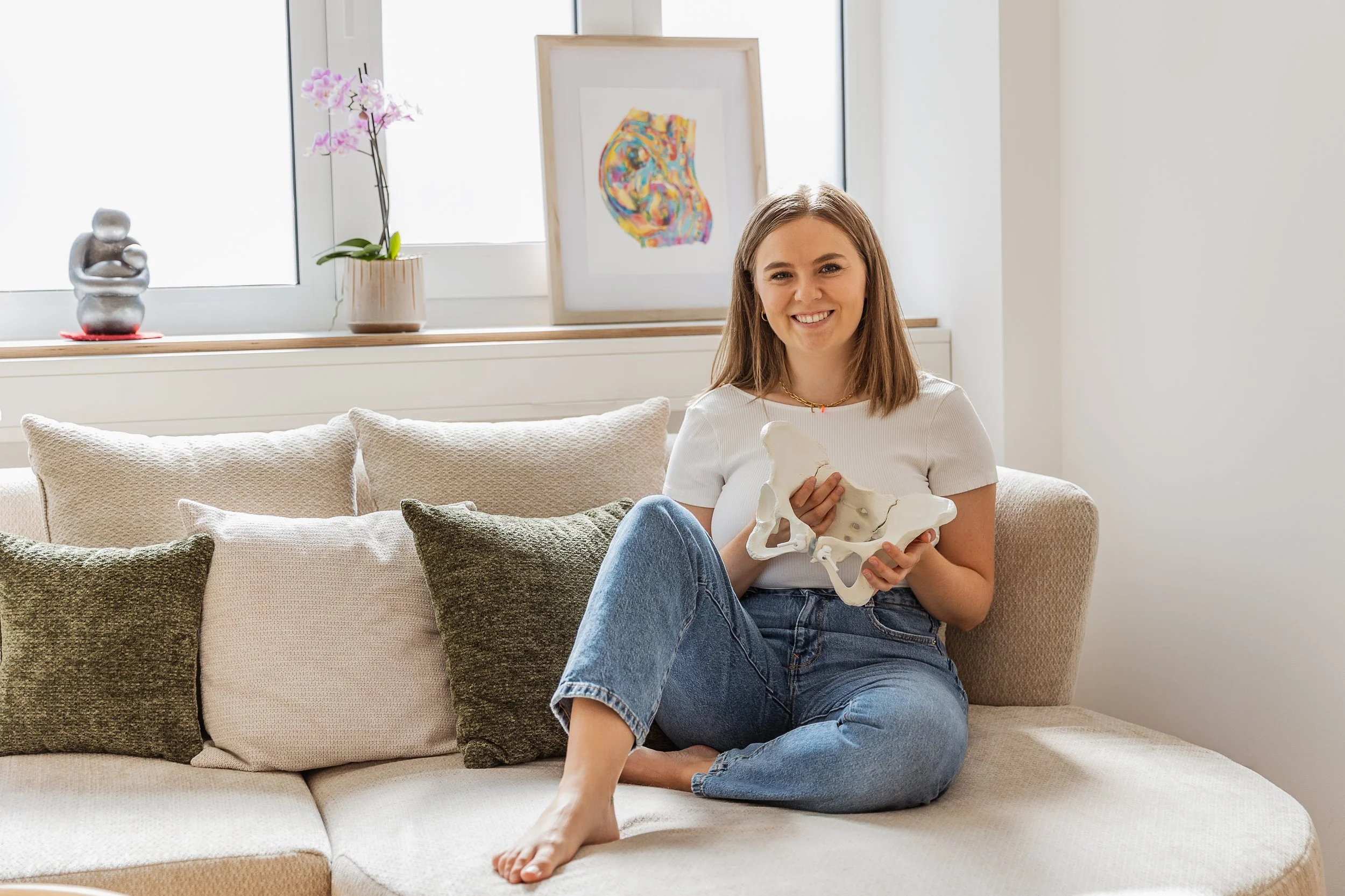 Eine junge Frau sitzt entspannt auf einem Sofa, hält ein menschliches Beckenmodell und lächelt in die Kamera, im Hintergrund sind eine Blume und bunte Kunst an der Wand.