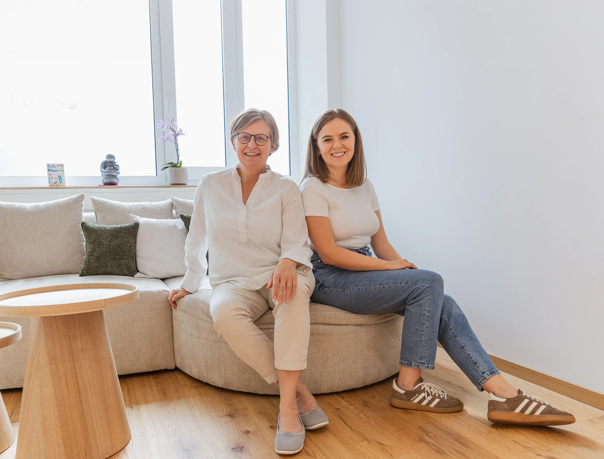 Zwei Frauen sitzen auf einer Couch in einem hellen Raum, lachen und schauen in die Kamera. Im Hintergrund sind große Fenster, eine Pflanze und eine kleine Karte im Fenster.