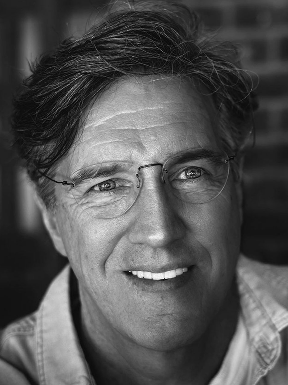 Black and white close-up photo of a smiling middle-aged man wearing glasses, with slightly tousled hair, in front of a brick wall.
