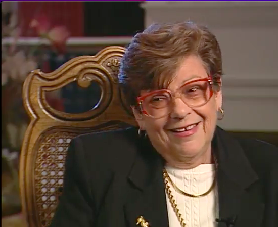 Older woman with short brown hair, red glasses, and gold jewelry sitting in a wooden chair and smiling.