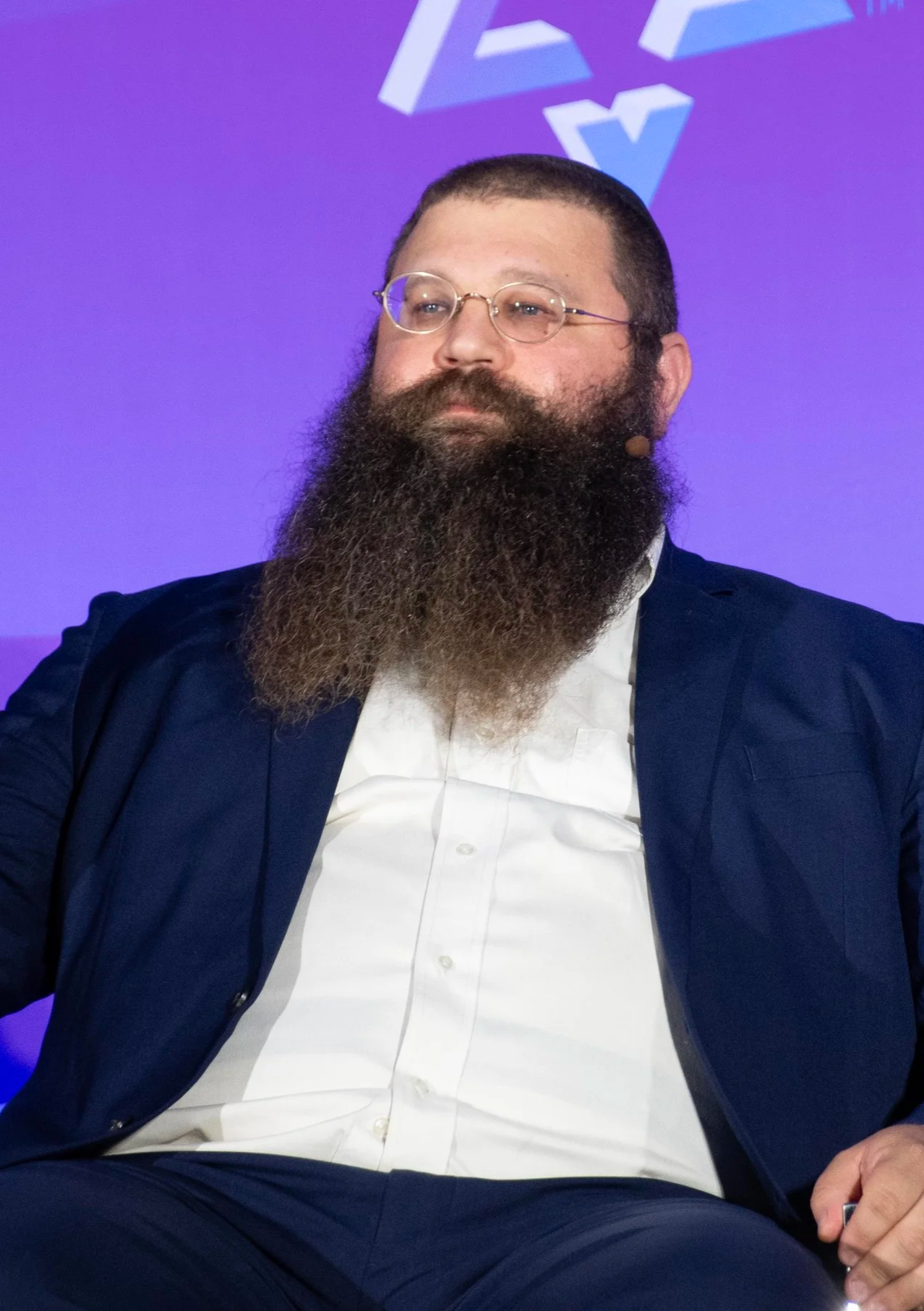 A man with glasses and a full beard wears a dark blazer and white shirt, sitting with a neutral expression against a purple background with abstract white shapes.