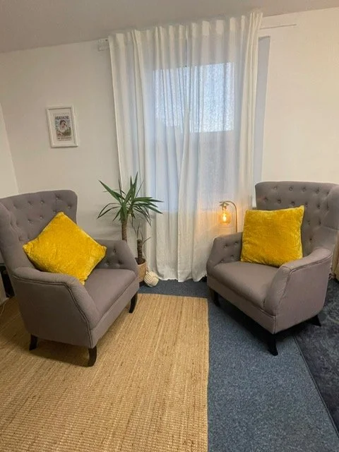 Two gray armchairs with yellow pillows facing each other in a room with a window, white curtains, a potted plant, and a small side table with a lamp. Picture of the counselling room