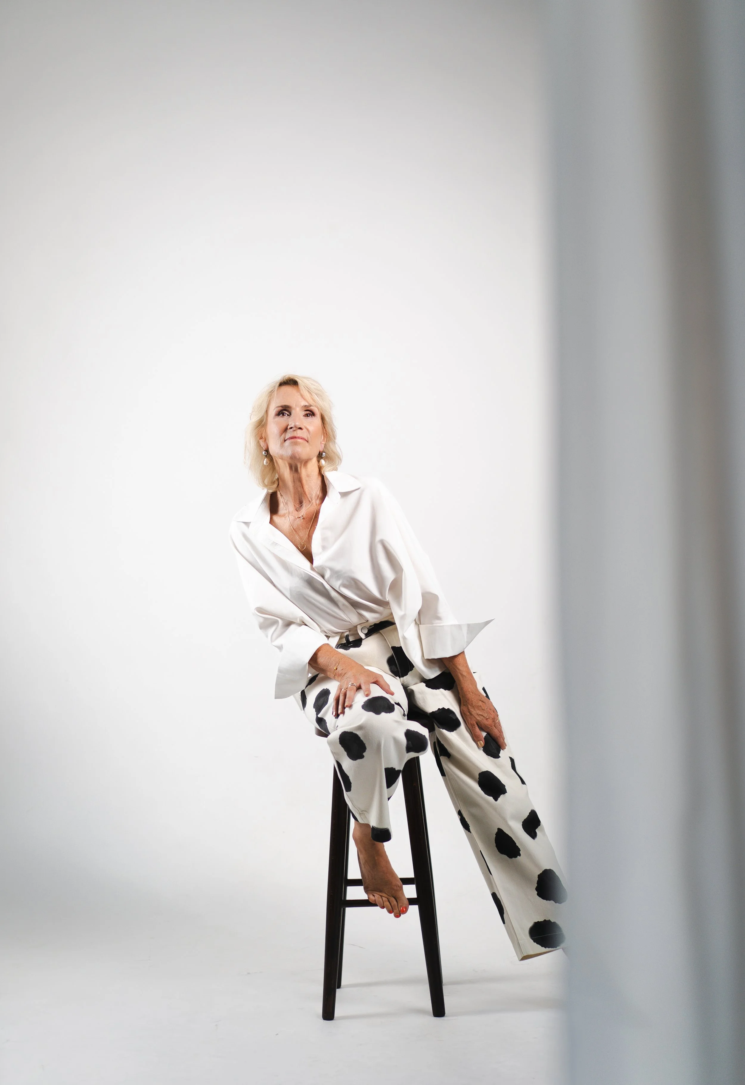 Een volwassen blonde vrouw zit op een zwarte kruk tegen een witte achtergrond. Ze draagt een witte blouse en een witte broek met grote zwarte stippen.