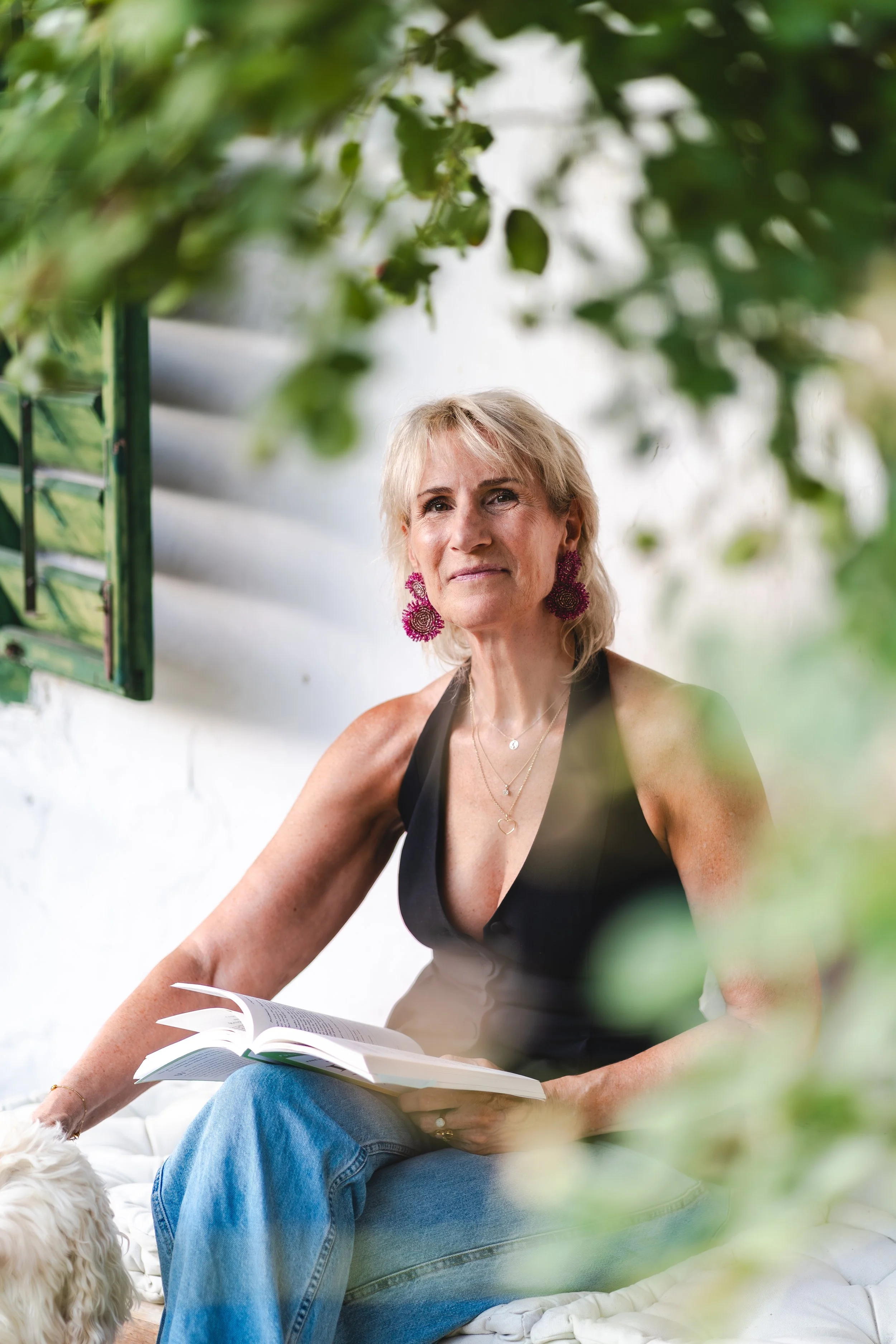 Een vrouw met kort blond haar, grote roze oorbellen en een zwarte top met een diepe halslijn zit op een bed of bank en leest een boek. Groene bladeren of planten bedekken een deel van het beeld.