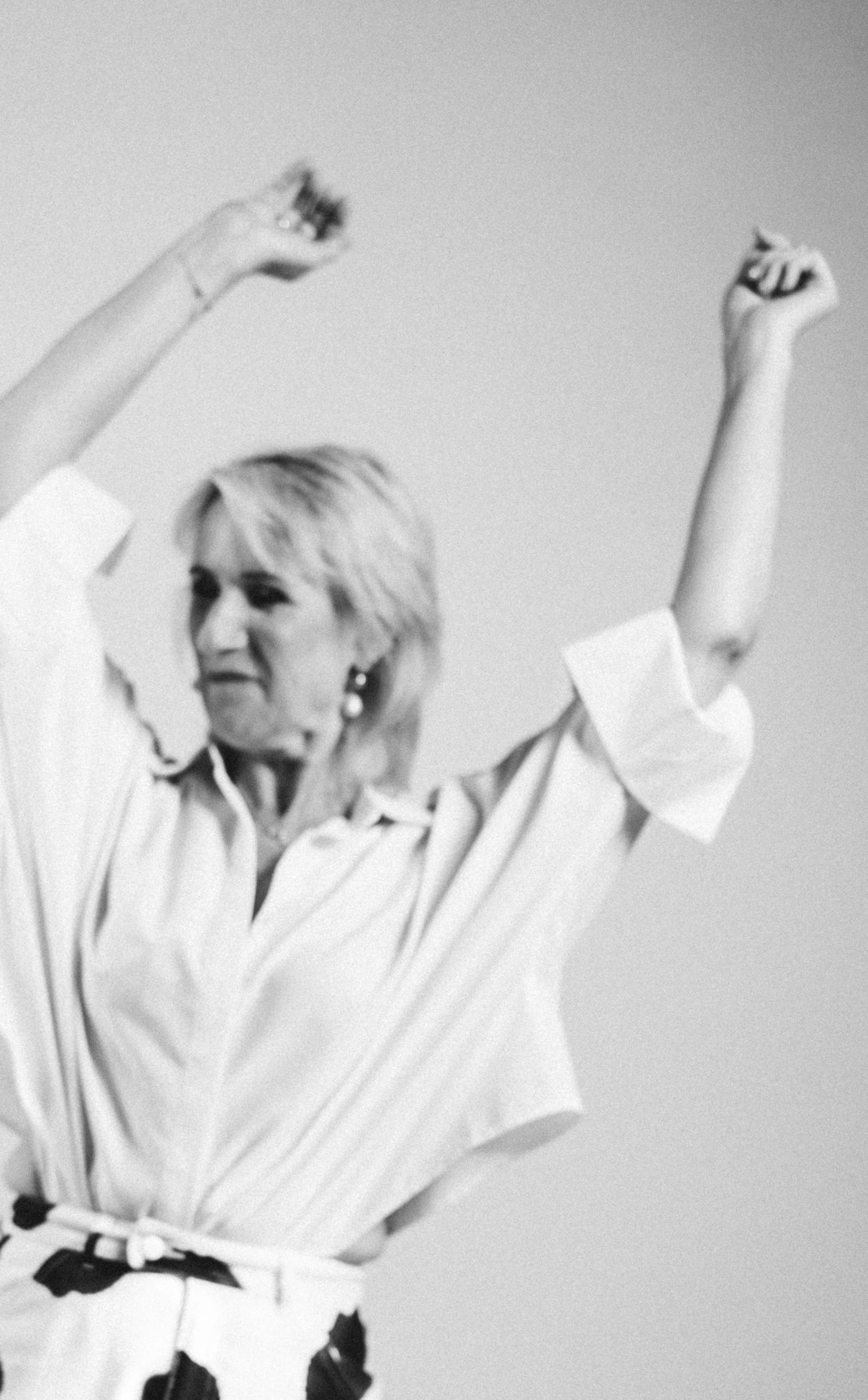 Een zwart-witfoto van een blonde, volwassen vrouw met kort haar, die feestelijk haar armen omhoog houdt. Ze draagt een witte blouse met zwarte patronen tegen een effen achtergrond.