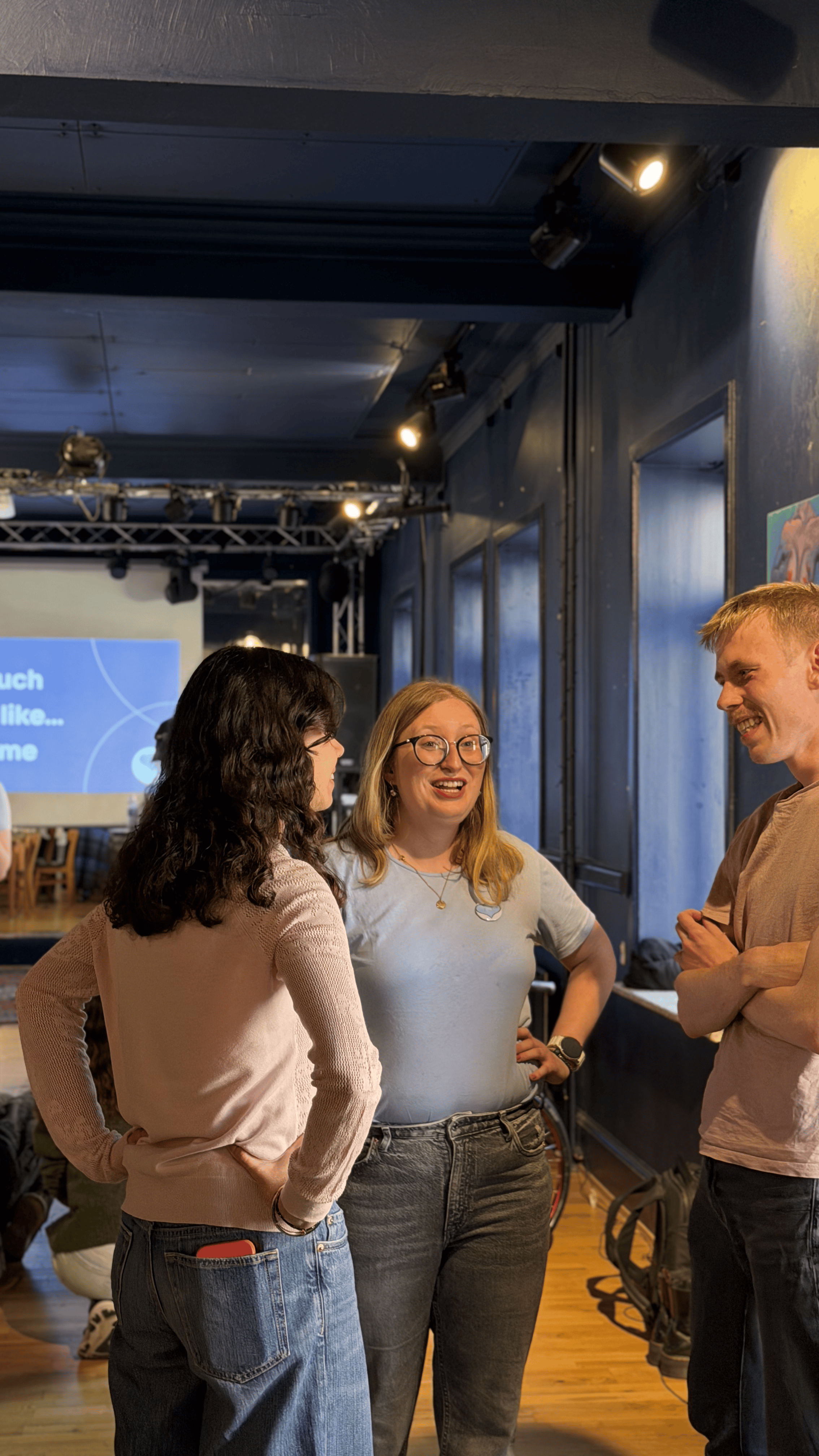 Three women and one man engaged in conversation in a dimly lit indoor space with windows and stage lighting. A presentation screen is visible in the background.