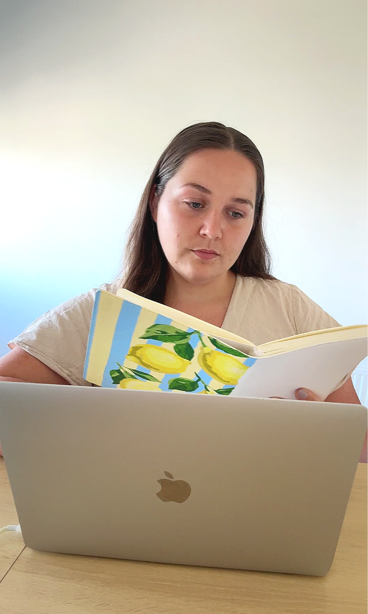 Une jeune femme avec des cheveux bruns, portant un t-shirt beige, regarde un livre avec des illustrations de citrons et de feuilles, posé sur un ordinateur portable gris avec une pomme gravée, sur une table en bois, dans un environnement intérieur
