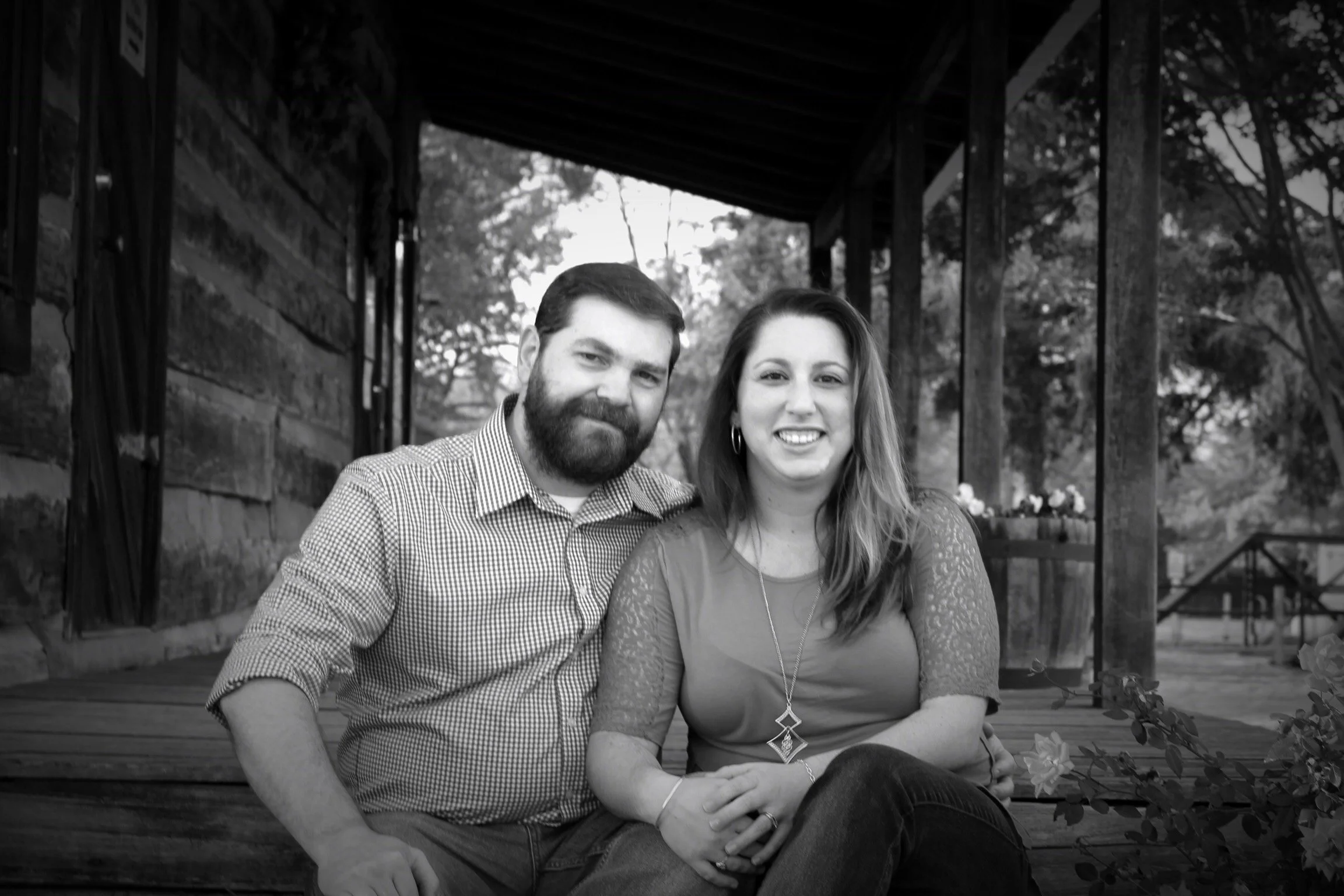 A black and white photo of a smiling couple sitting on a wooden bench outdoors, with trees and a patio structure in the background.