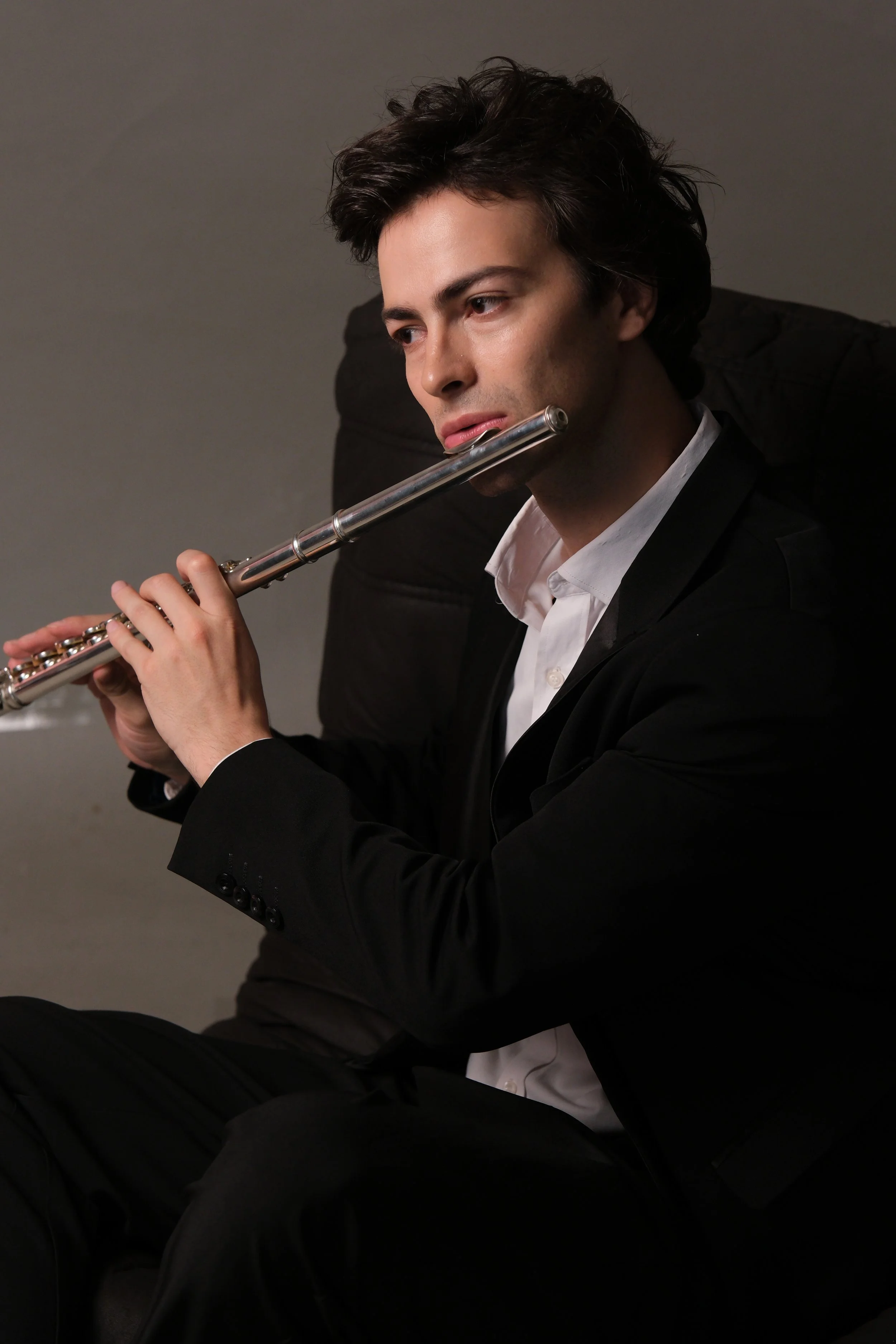 A young man in a black suit and white shirt playing a silver flute.