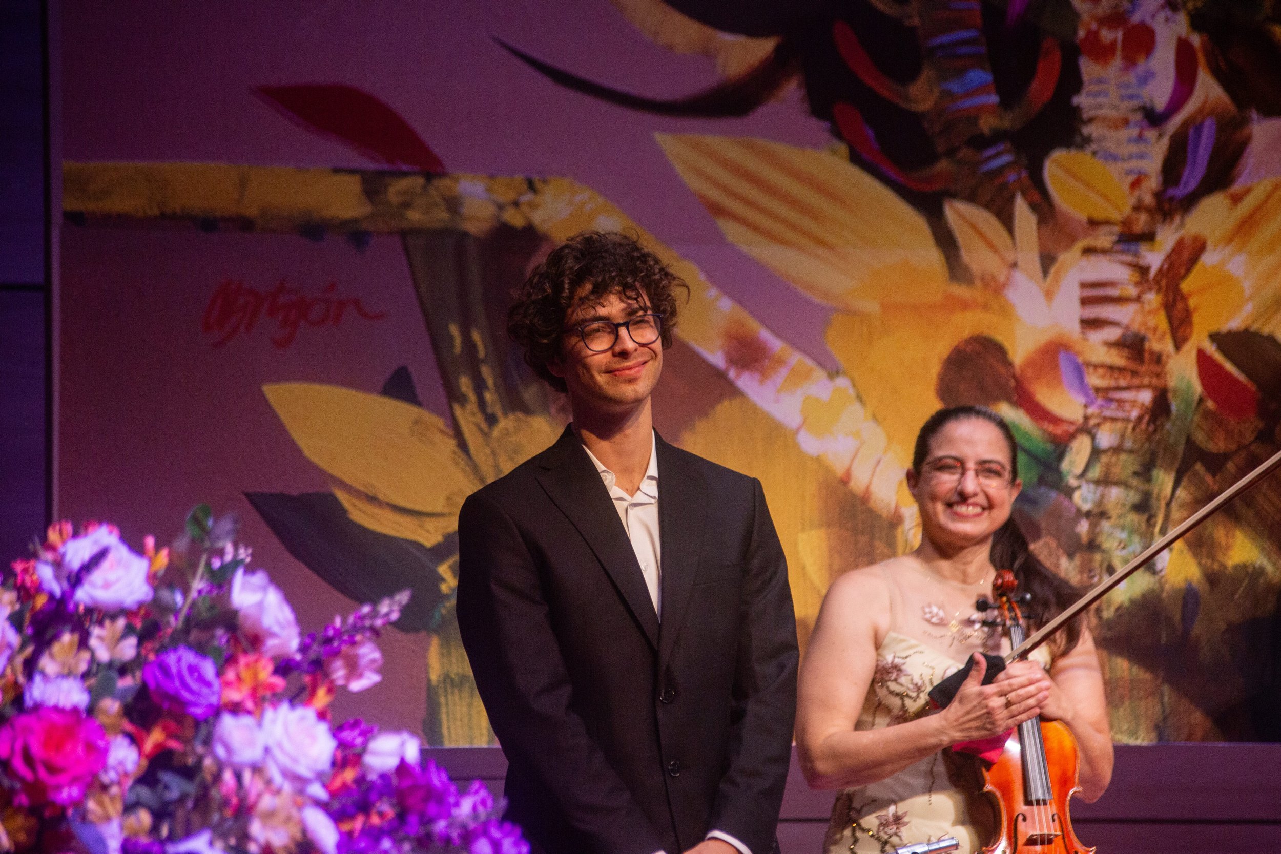 A man with curly hair, glasses, wearing a black suit and white shirt, smiling at an event. Next to him is a woman holding a violin and bow, wearing a sleeveless dress with floral embroidery, smiling. The background features colorful abstract artwork and purple flowers.