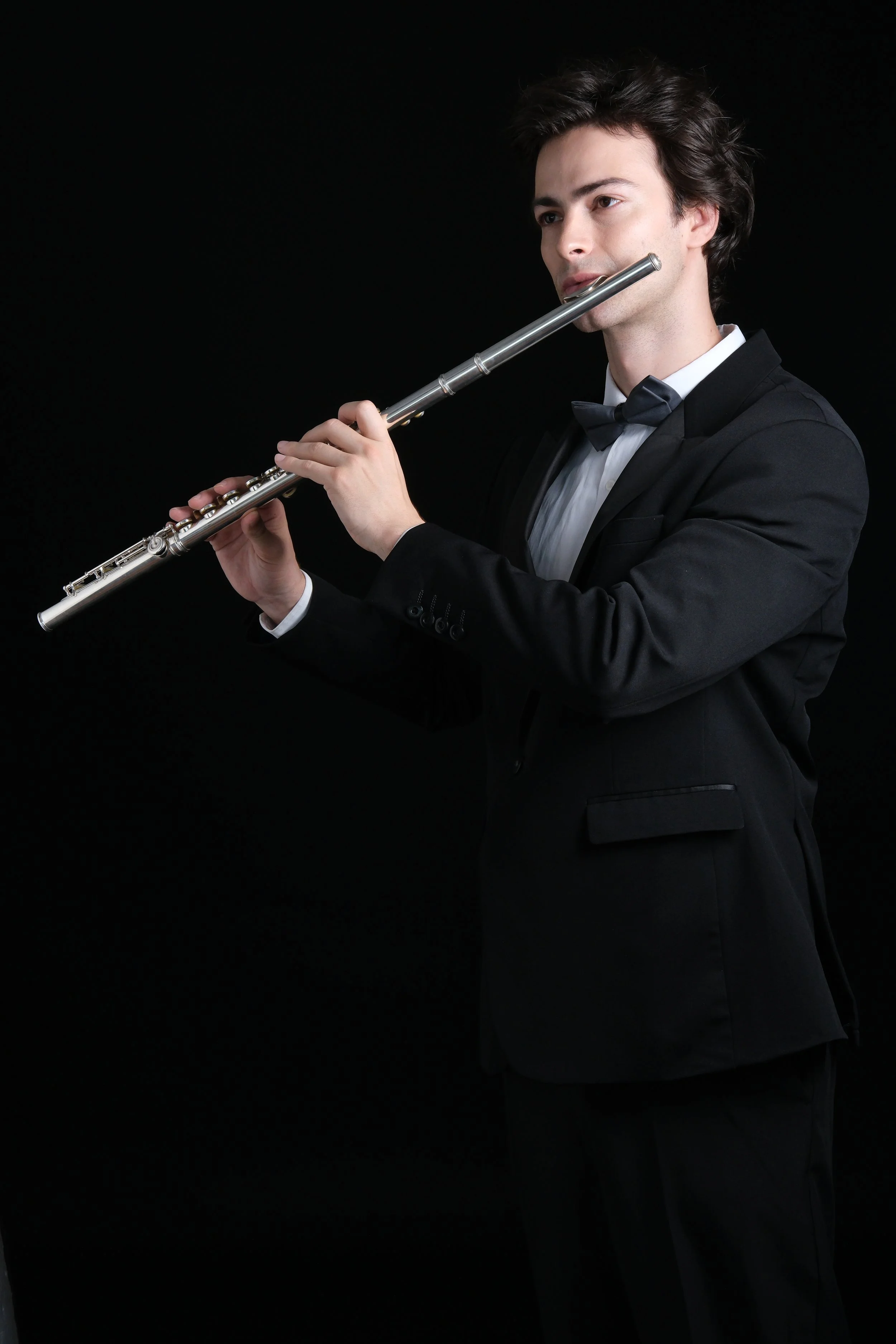 A man dressed in a black tuxedo with a bow tie, playing a silver flute against a black background.