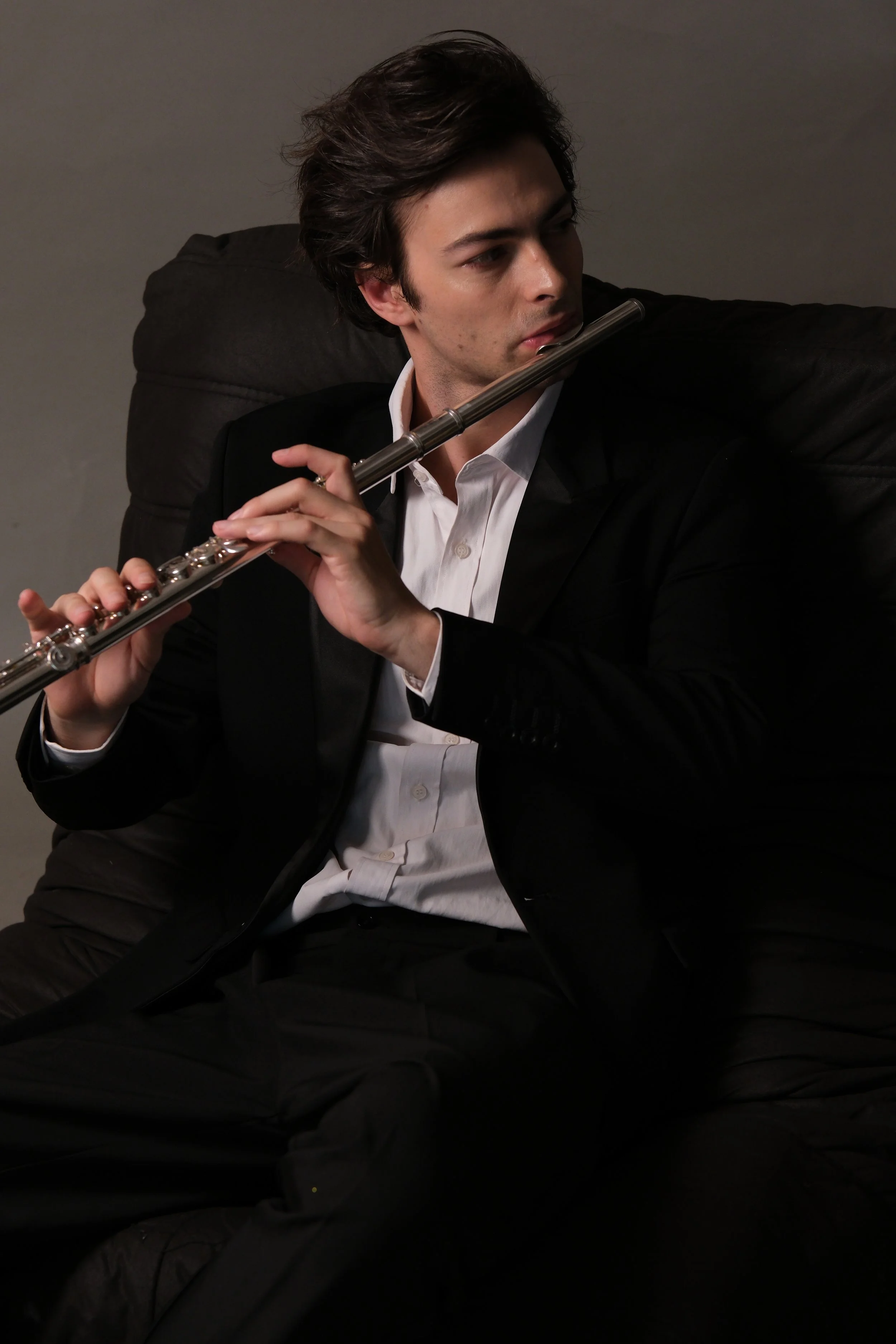 A young man in a black suit and white shirt sitting on a black couch, holding a silver flute near his face, and looking down thoughtfully.