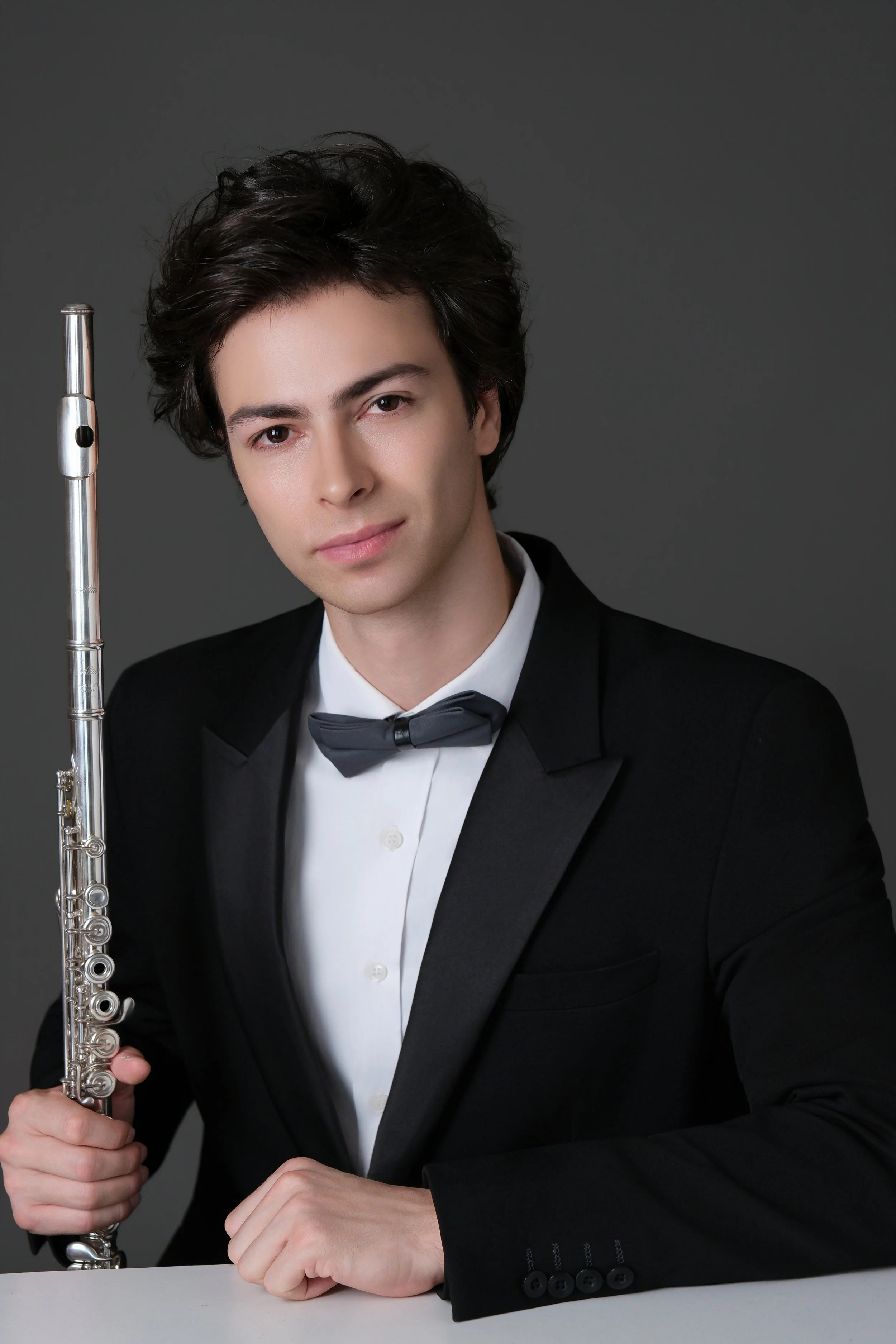 A young man with dark, wavy hair, dressed in a tuxedo, holding a flute, sitting at a white table against a dark gray background.