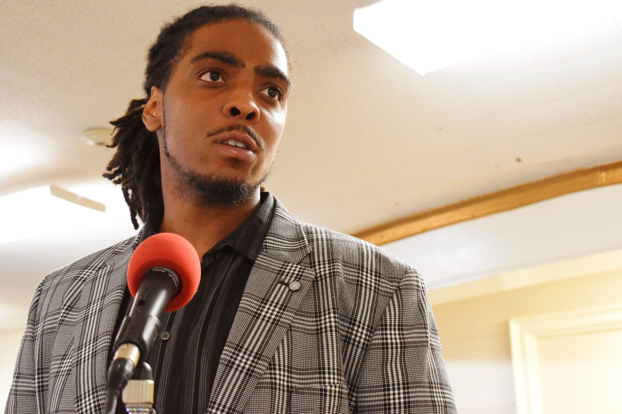 A man with dreadlocks and a goatee speaking into a red microphone, wearing a checkered blazer and a black shirt, indoors with beige walls and wood accents.