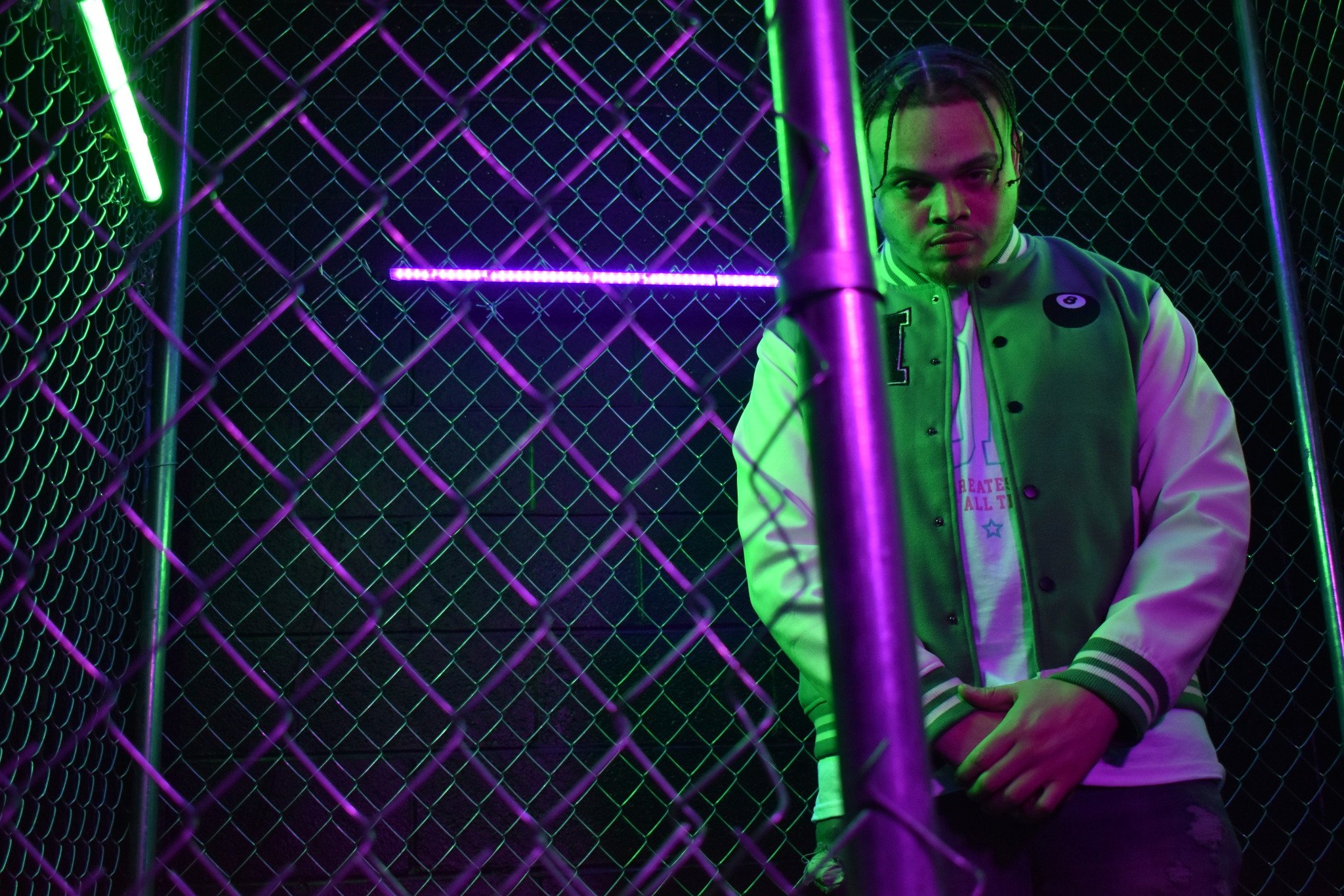 A young man with braided hair wearing a varsity jacket standing behind a chain-link fence illuminated by neon purple and green lights.