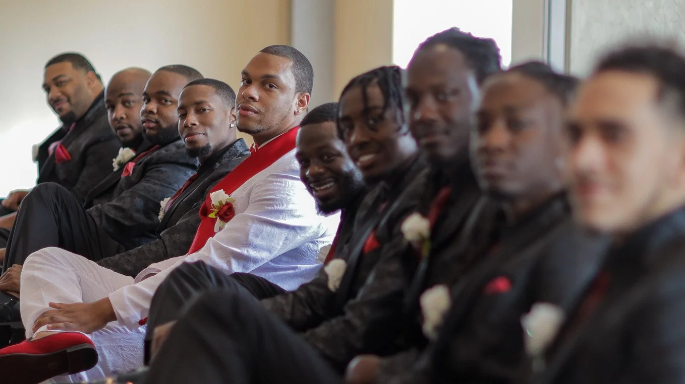 A group of men sitting in a row, dressed formally in suits, at a wedding or formal event.