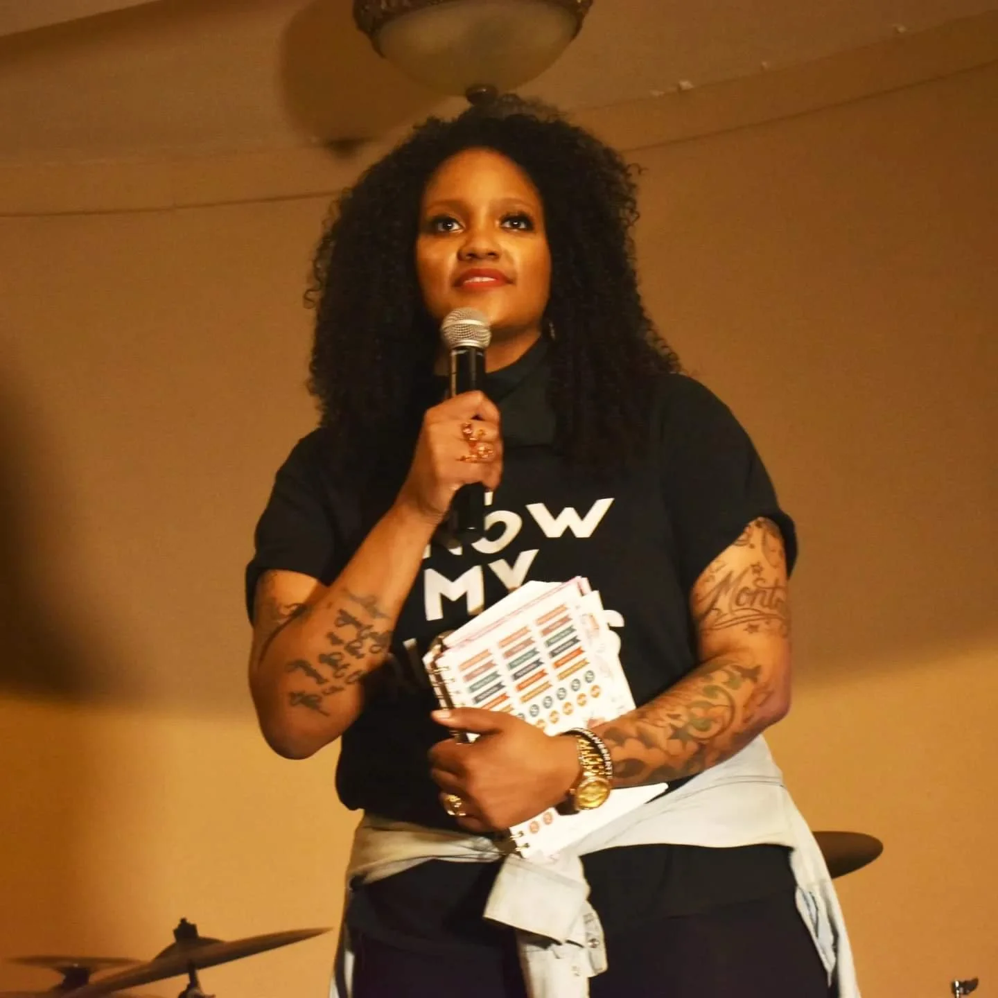 A woman with curly hair holding a microphone and some papers, wearing a black t-shirt, standing in a room with a beige wall and a ceiling fan.