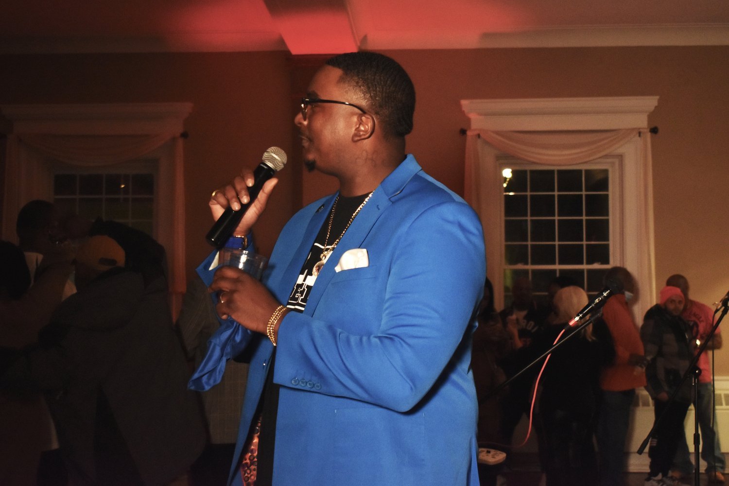A man wearing a bright blue blazer, glasses, and necklaces, holding a microphone and a drink, standing at an indoor event with people in the background.