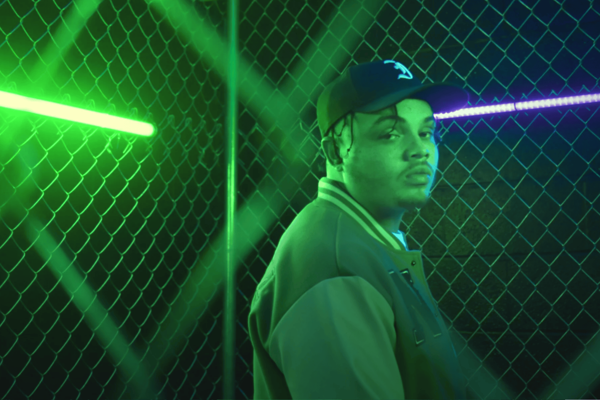 A young man with braided hair wearing a cap with a logo, standing near a chain-link fence illuminated by green and purple neon lights.