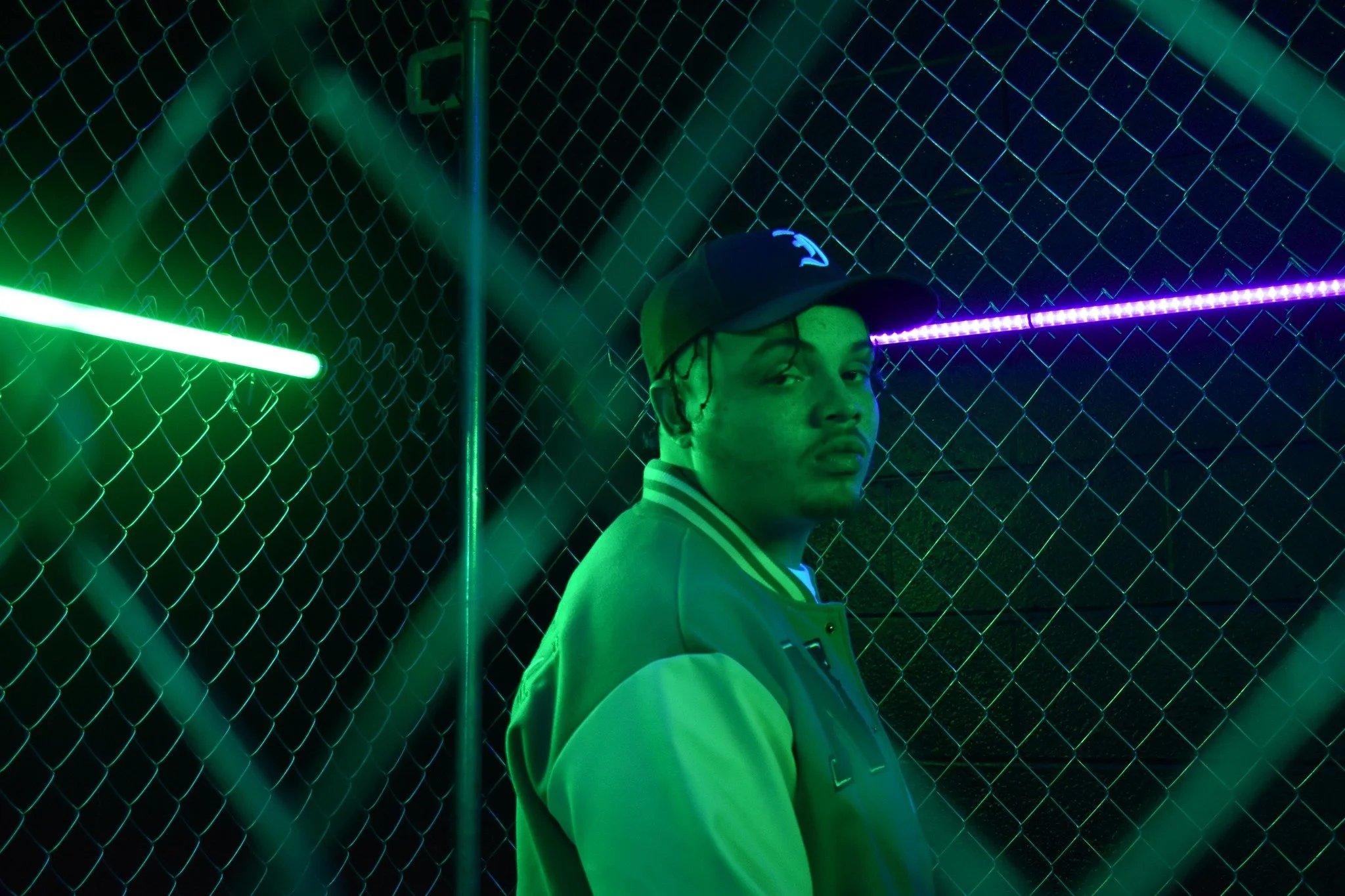 A young person wearing glasses and a baseball cap stands beside a chain-link fence illuminated with green and purple neon lights at night.
