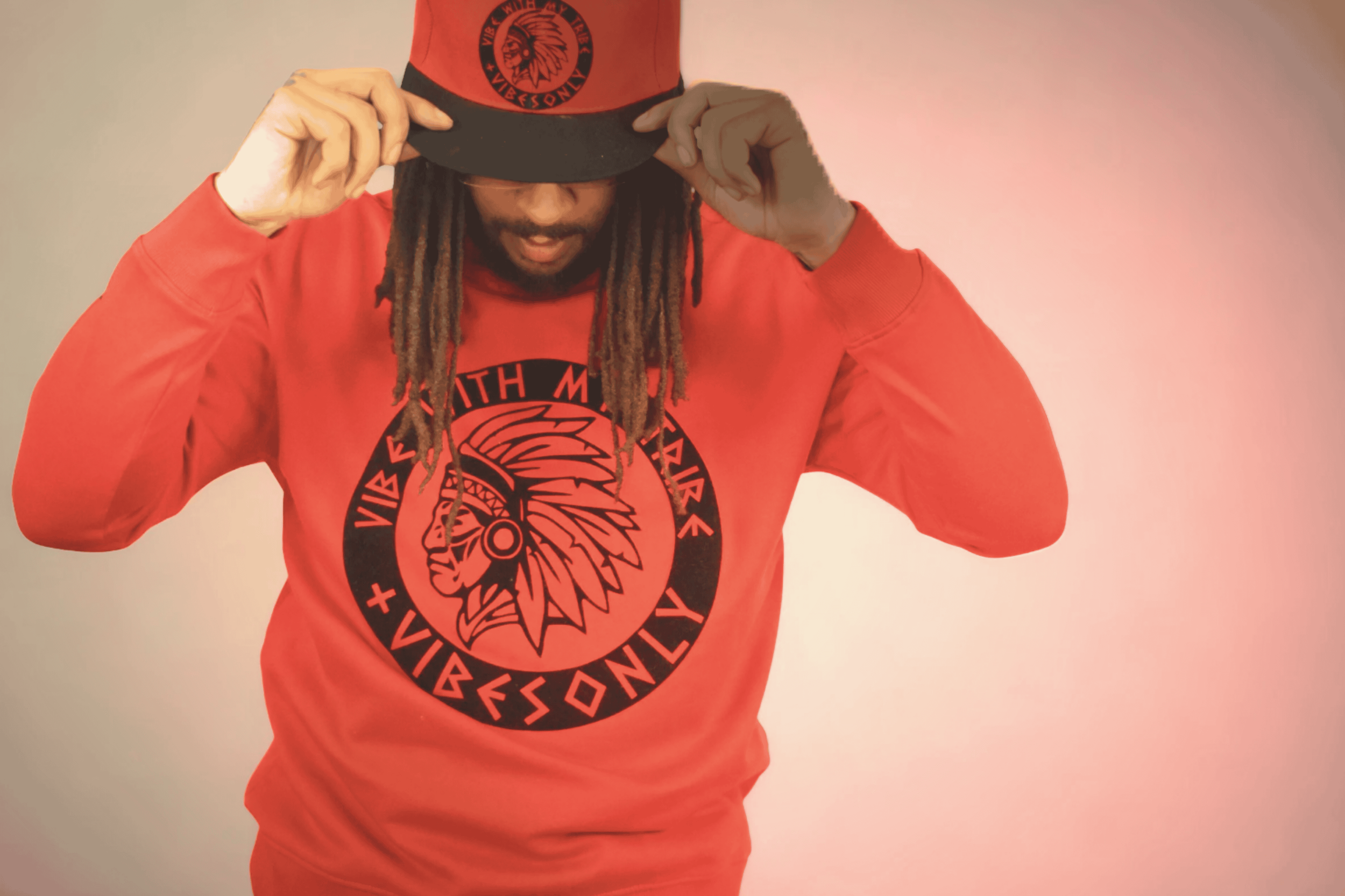 A person wearing a red sweatshirt and a red and black cap, adjusting the cap by holding its brim with both hands. The sweatshirt and cap display a circular logo with a stylized Native American headdress and the text 'VIBE' and other words, along with