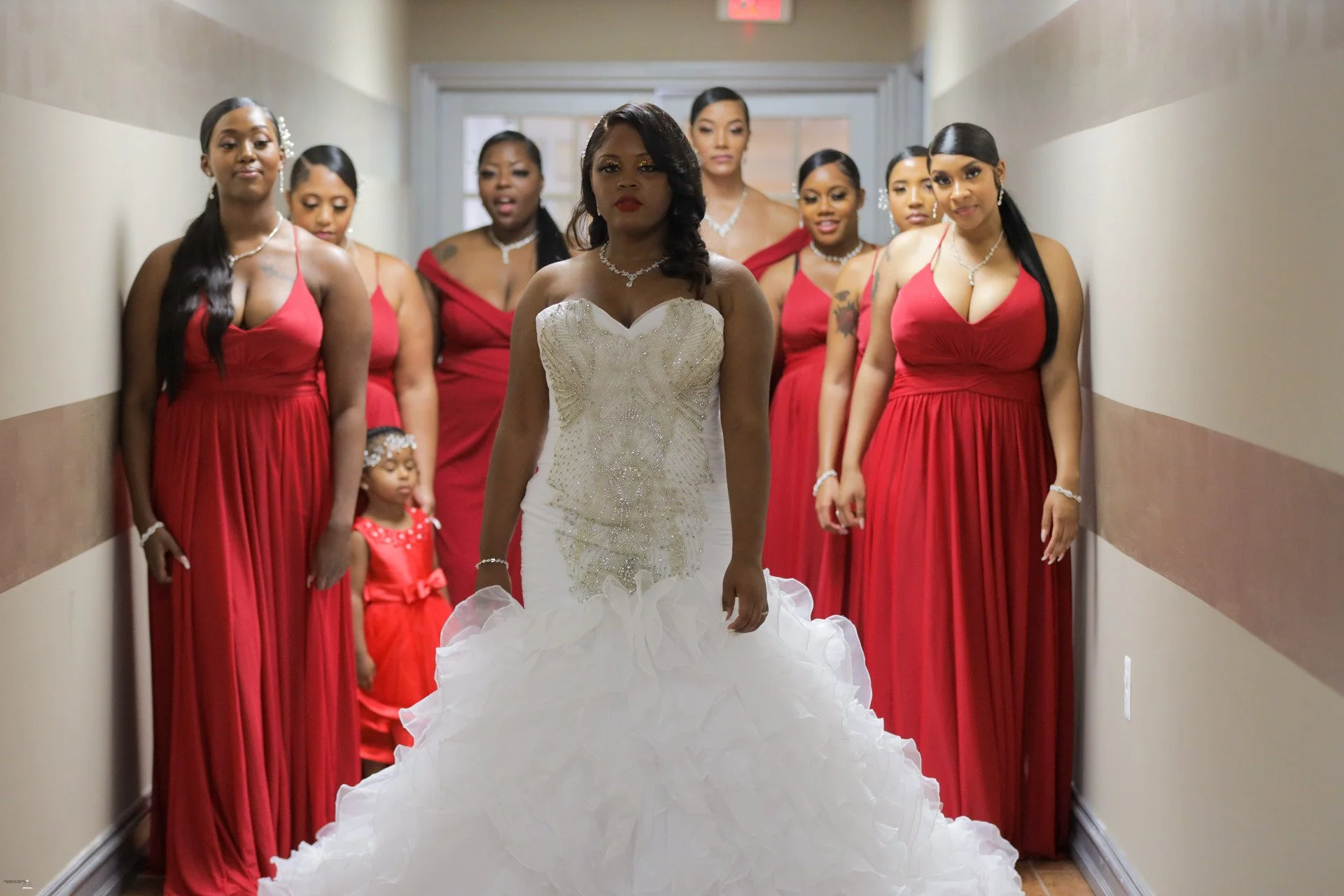 A bride in a white wedding gown walking down the hallway with eight bridesmaids in red dresses and a young flower girl in a red dress following behind.