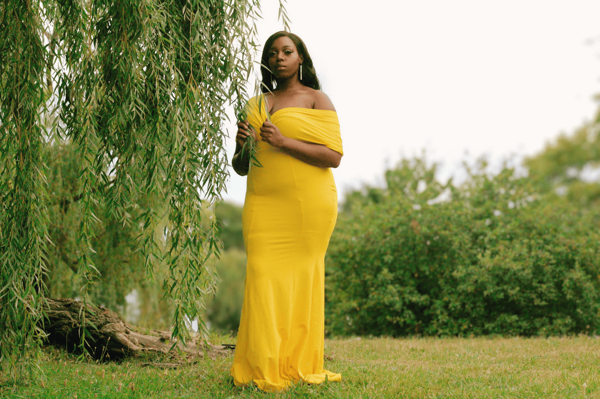 A woman in a yellow dress standing outdoors near a tree with drooping branches and green foliage.