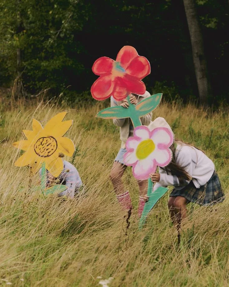Children holding paper flower cutouts in a grassy field with trees in the background.