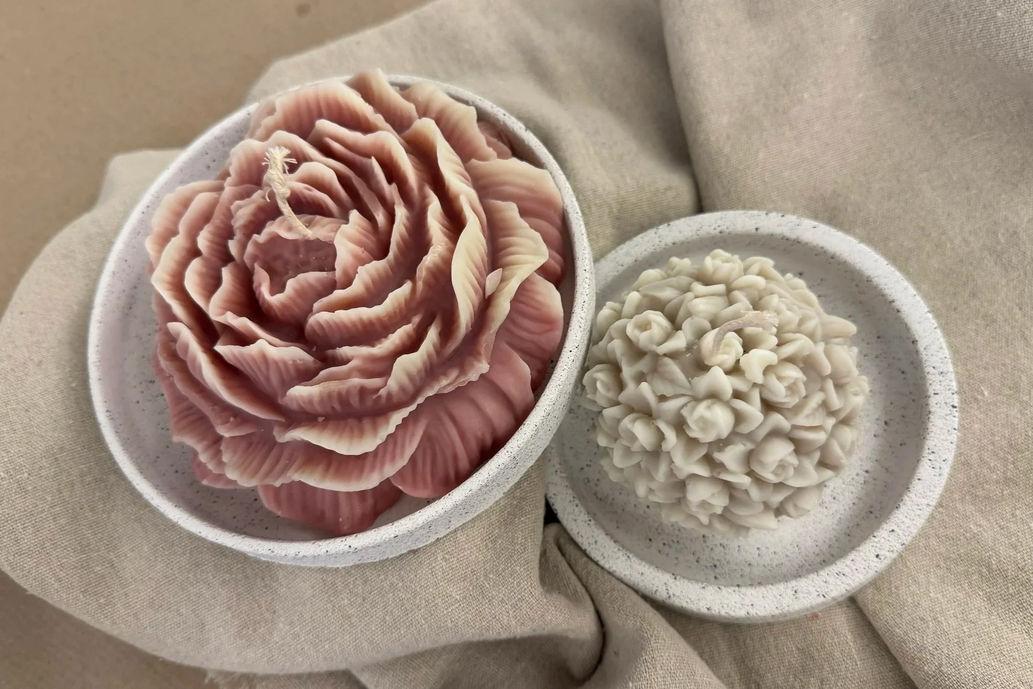 Zwei ovale Schälchen mit strukturierter Oberfläche und Seifenstücken in Form einer Rose und eines Gehirns, auf beigem Stoff – THALIE DÉCO.