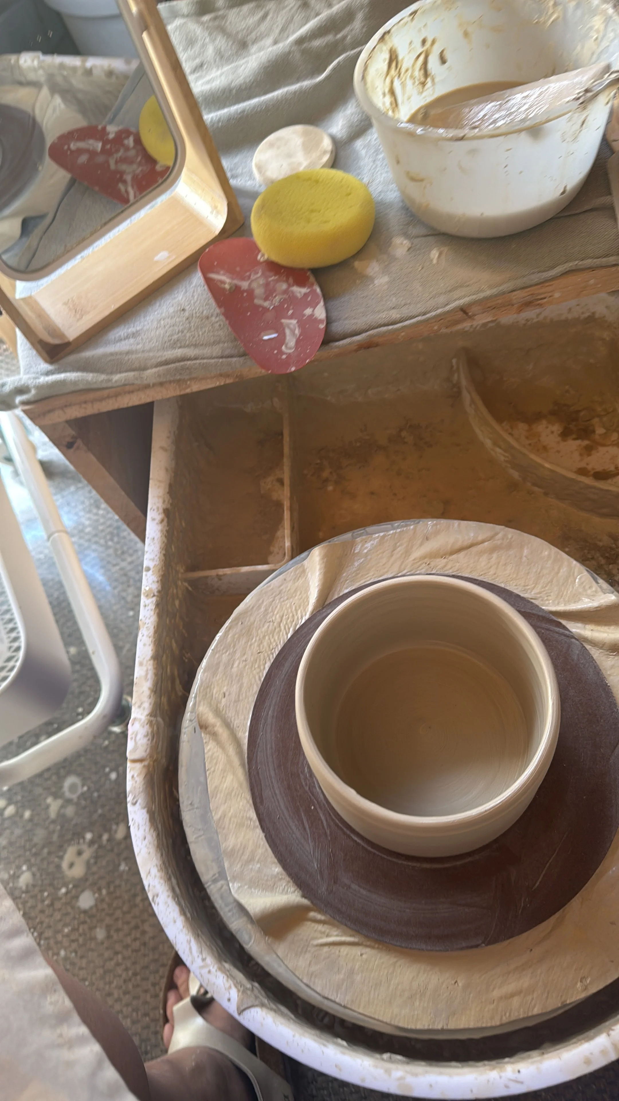 Töpferscheibe mit einer entstehenden Keramiktasse, umgeben von Werkzeugen, Schwamm und einer Wasserschale – aktive Keramikarbeit bei THALIE DÉCO.