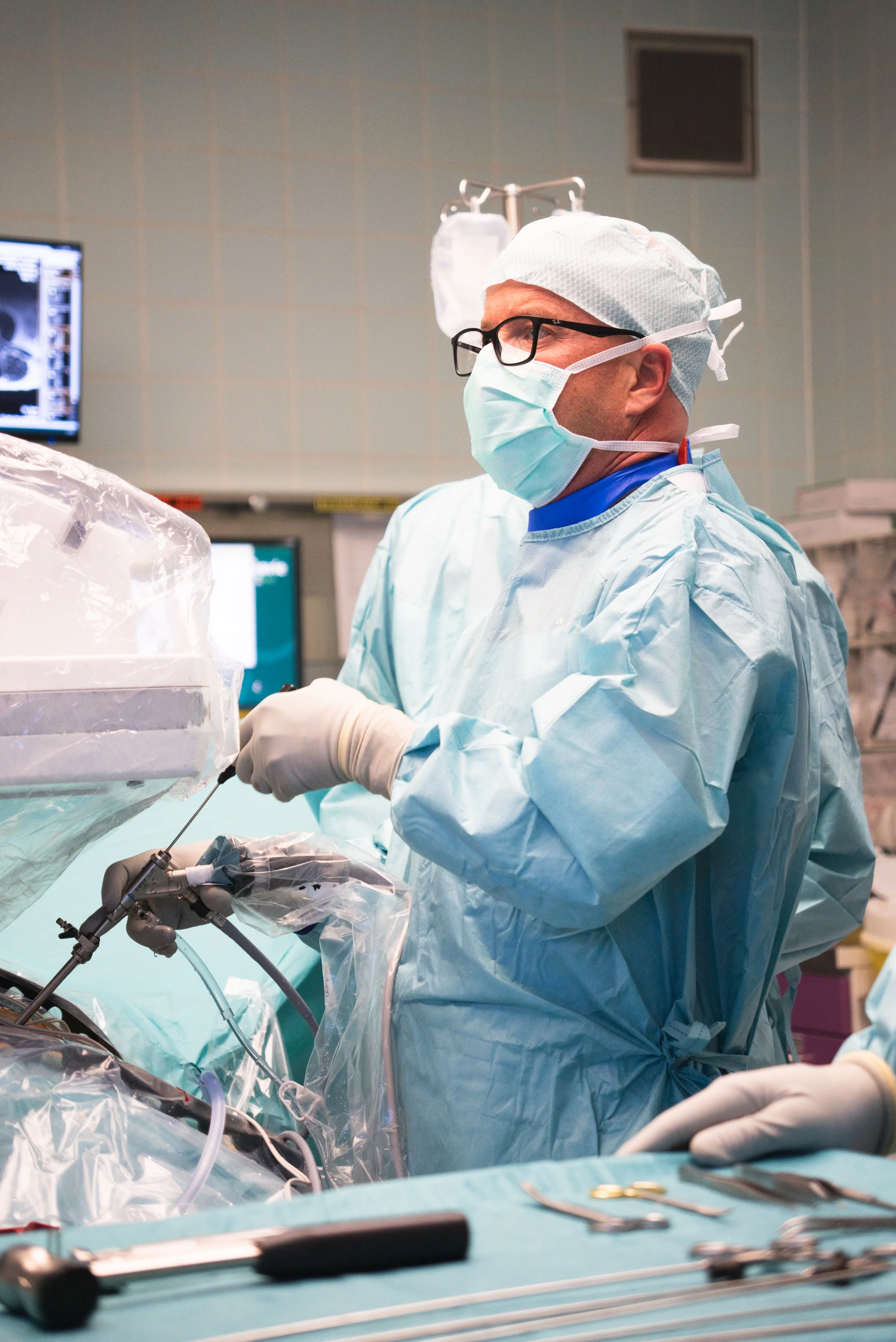 Ein Chirurg in blauer OP-Kleidung, Maske und Brille arbeitet während einer Operation im Operationssaal. Es gibt medizinische Geräte und Monitore im Hintergrund.