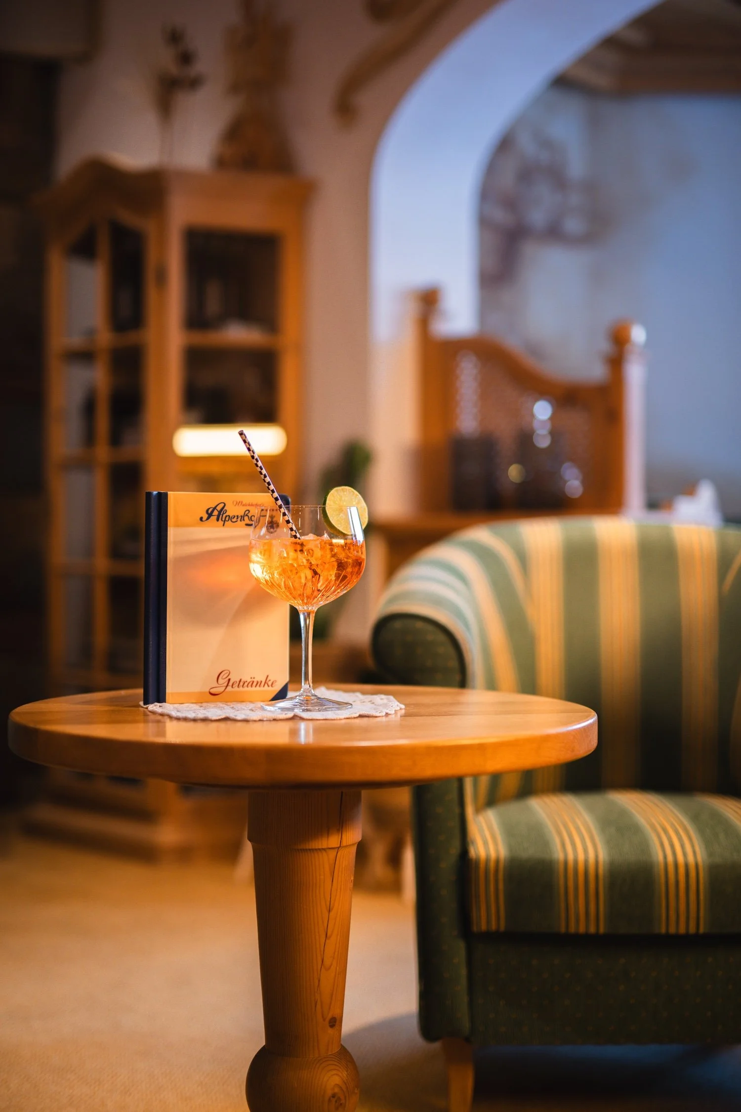 Ein Cocktail mit Orangenscheibe und einem gestreiften Strohhalm auf einem Holz-Couchtisch in einem gemütlichen Raum.
