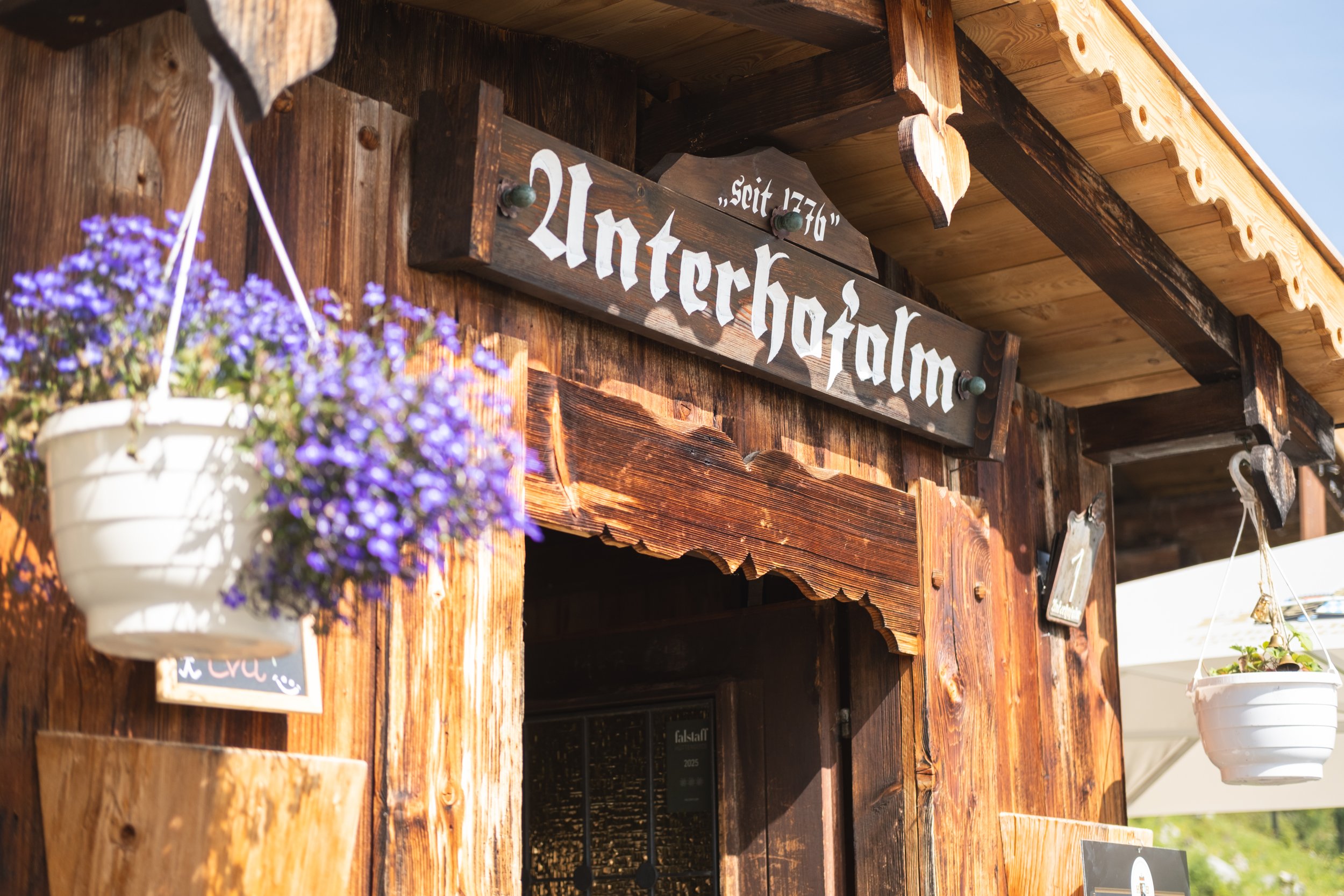 Das Bild zeigt eine Holzhütte mit einem Schild, auf dem 'Sitz 17/40' geschrieben steht, und zwei hängende Blumenampeln mit lila Blumen vor der Hütte.